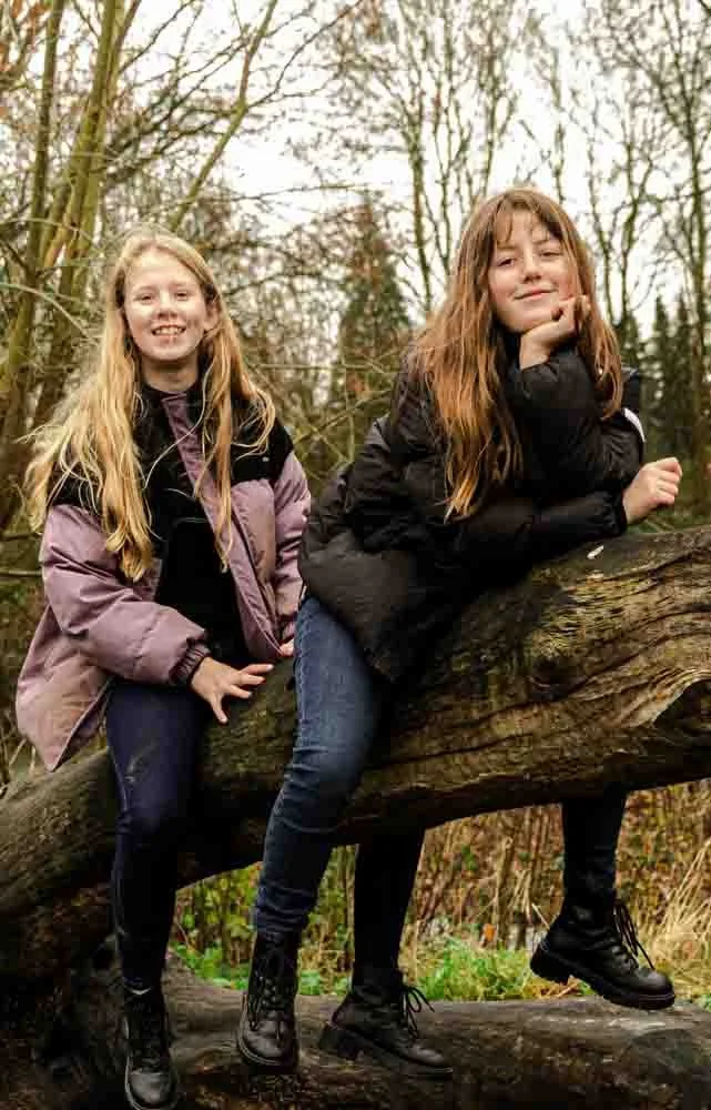 Twee jonge meisjes zitten op een omgevallen boom in een bos, lachend en poserend voor de camera.