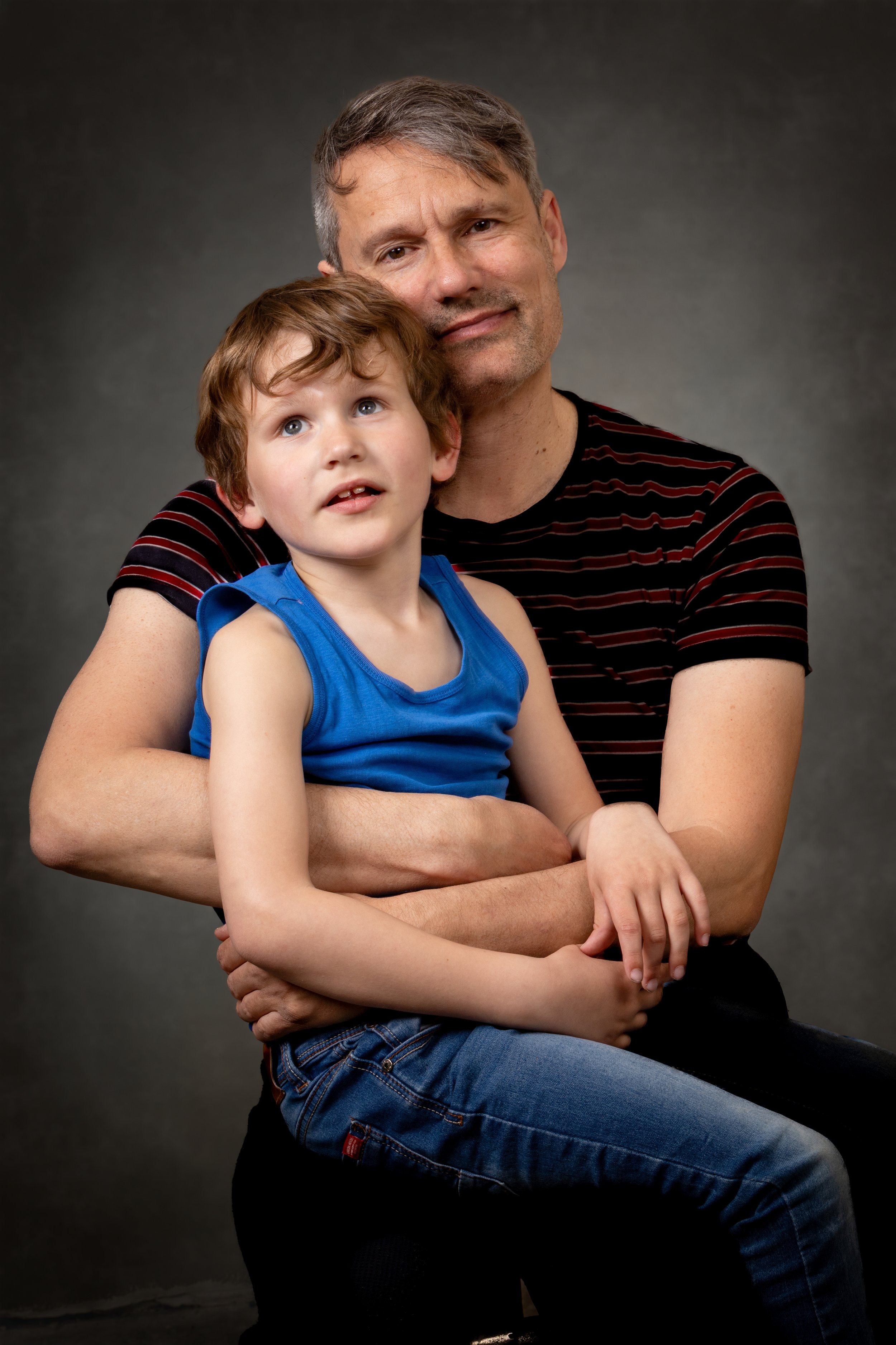 A man and a young boy sitting together, with the man holding the boy. The man has short grayish hair and is wearing a black shirt with red stripes. The boy has curly brown hair and is wearing a blue sleeveless shirt and jeans. Both are looking slightly upward, with a neutral to curious expression, against a plain gray background.