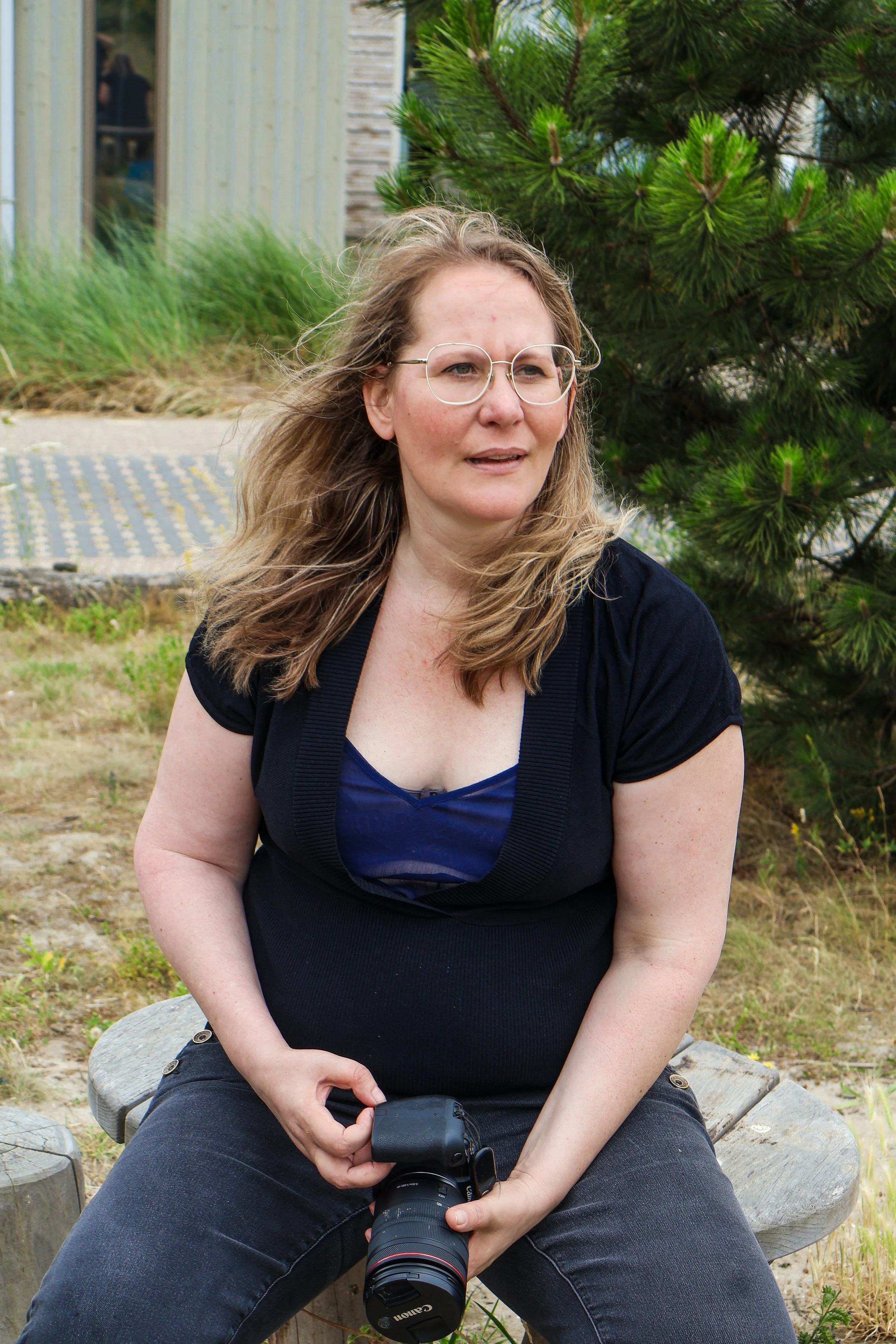 A woman sitting on a wooden bench outdoors, holding a Canon camera, with curly blonde hair, wearing glasses, a dark top, and jeans, near a tree with green foliage.