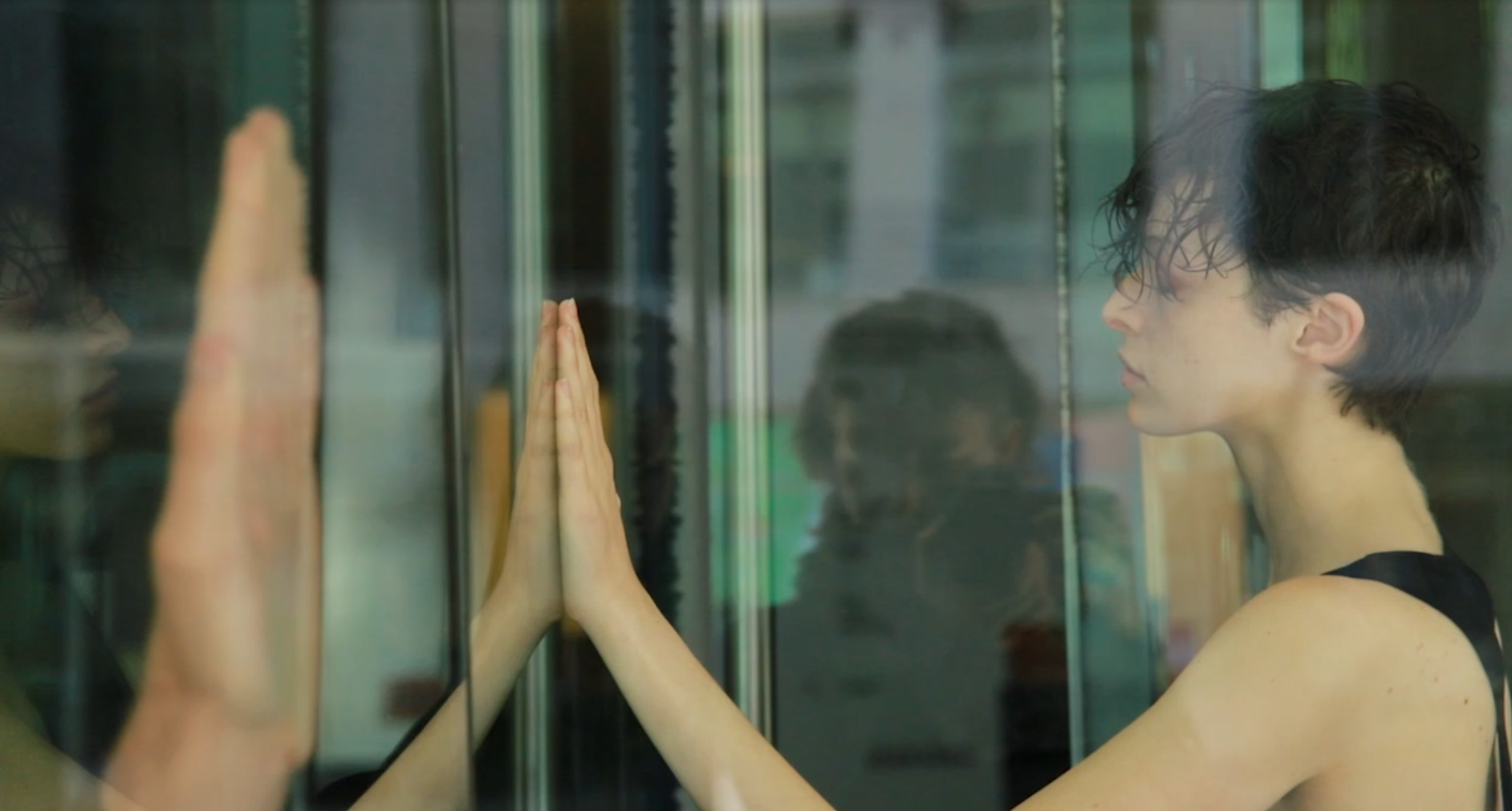 A person with short curly black hair and light skin is pressing their palms against a glass surface, touching their reflection.