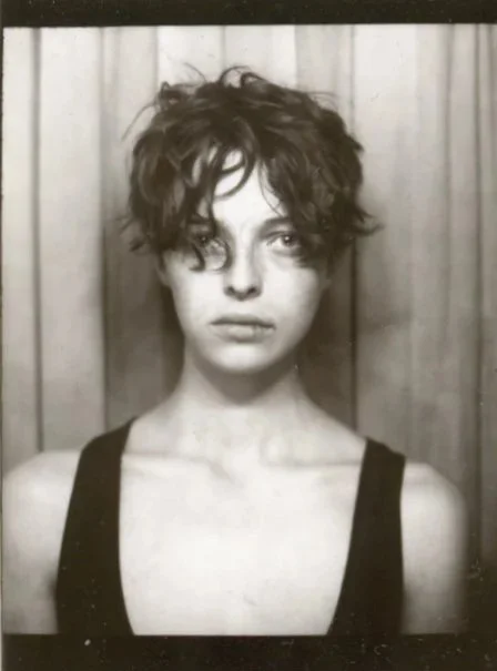 Black and white portrait of a young woman with short, curly hair, wearing a sleeveless top, standing in front of a wooden wall.