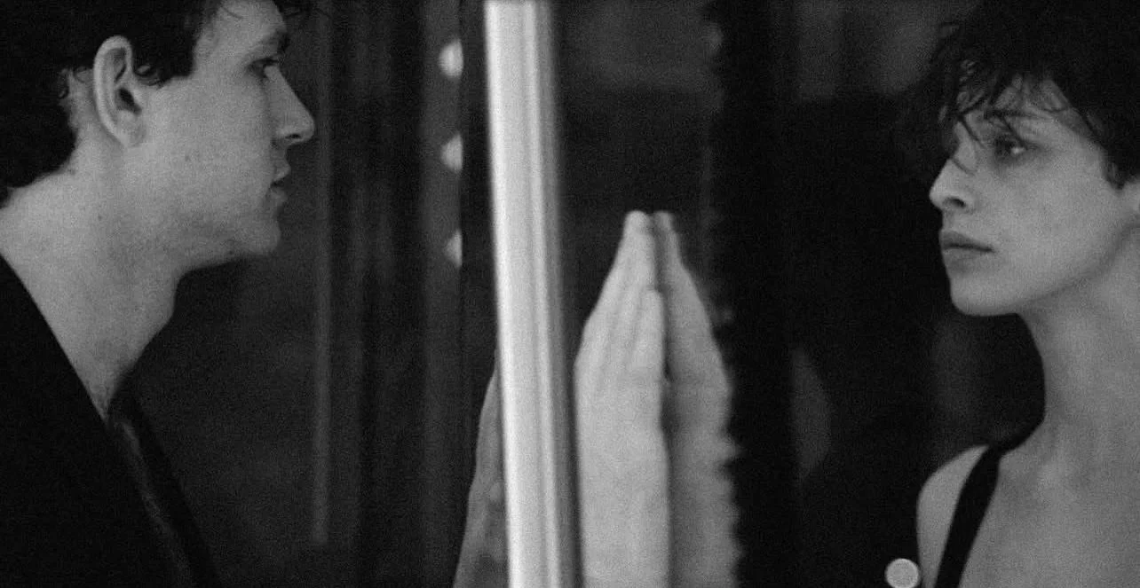 A black and white photo of a young man and woman facing each other with a glass between them, both touching the glass with their hands and looking into each other's eyes.