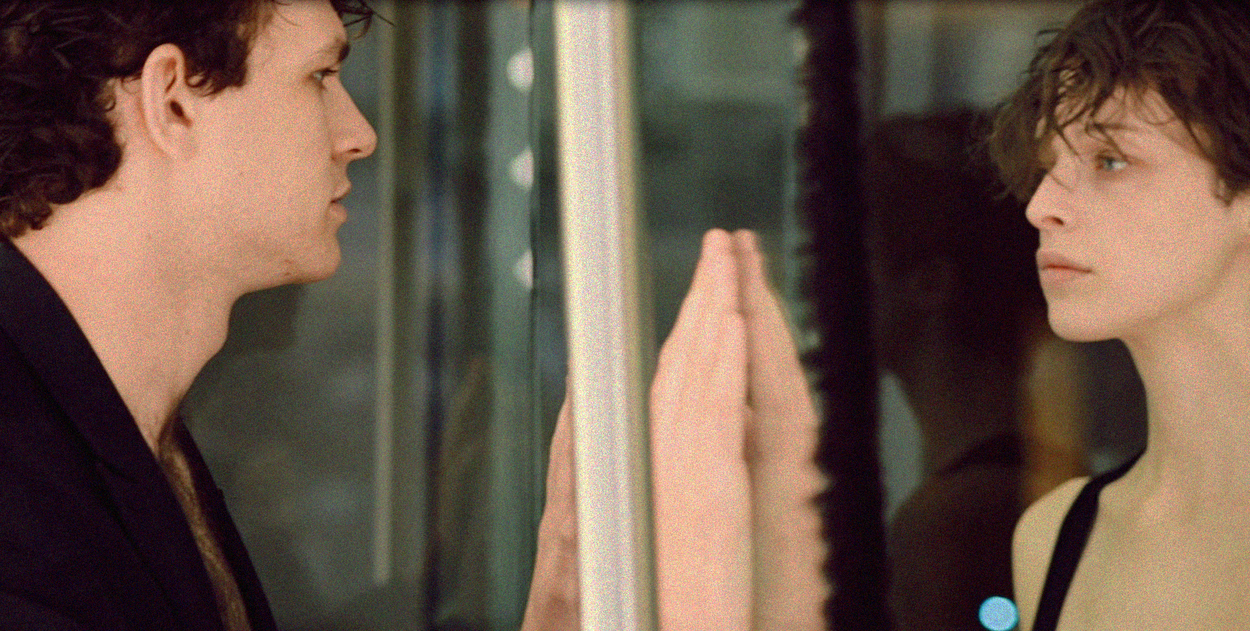 A young man and woman look at each other through a glass window, with their hands pressed against the glass.