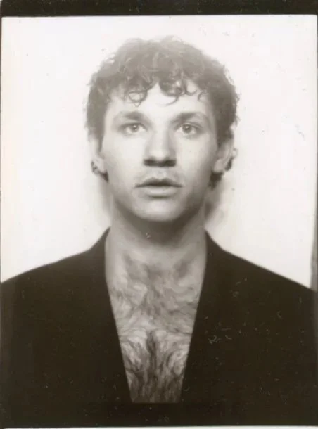 Black and white portrait of a young man with curly hair, open mouth, and wearing a dark jacket with an open shirt revealing chest hair.