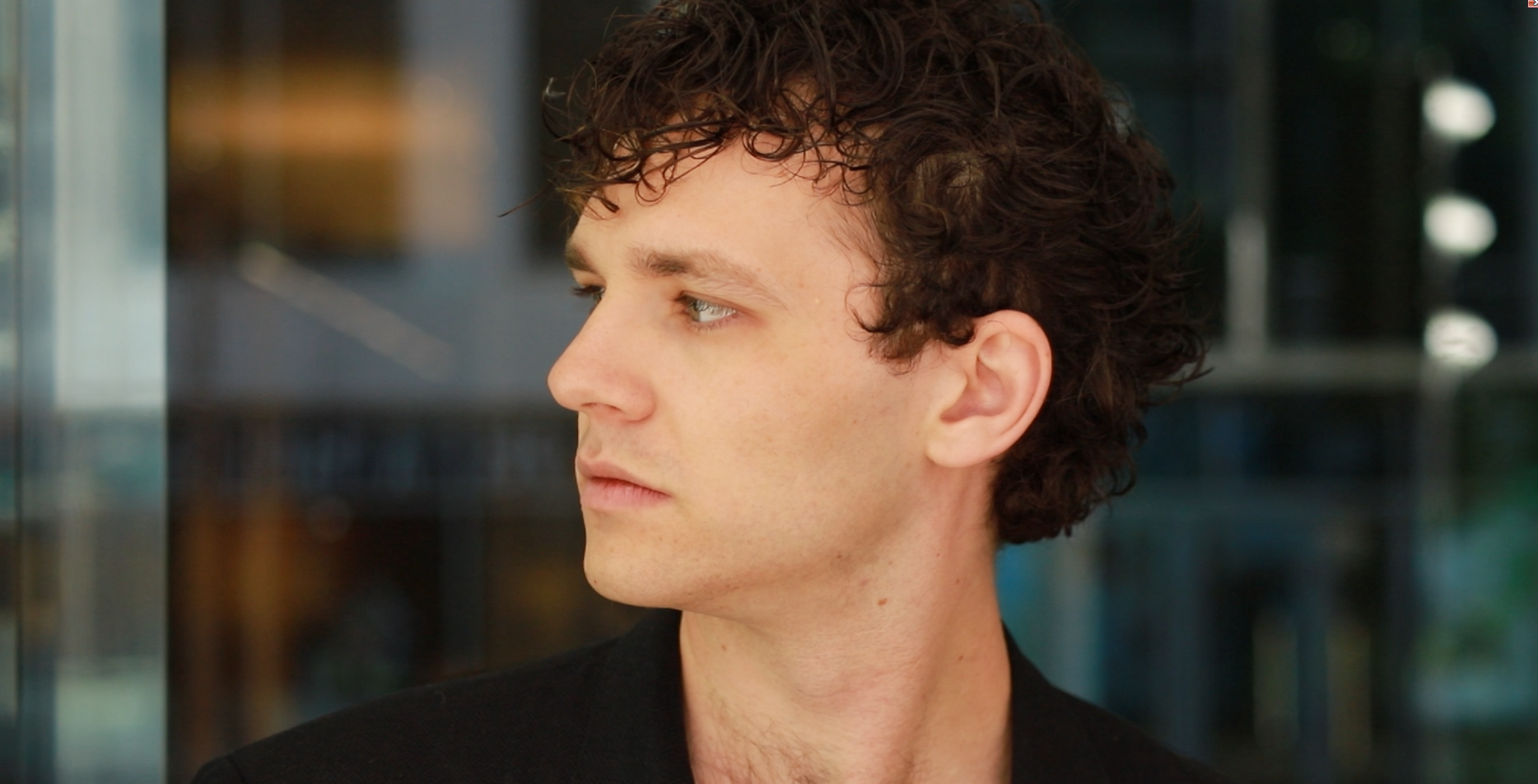 Side profile of a young man with curly brown hair, looking to the left. He is wearing a black shirt and standing indoors with a blurred background.
