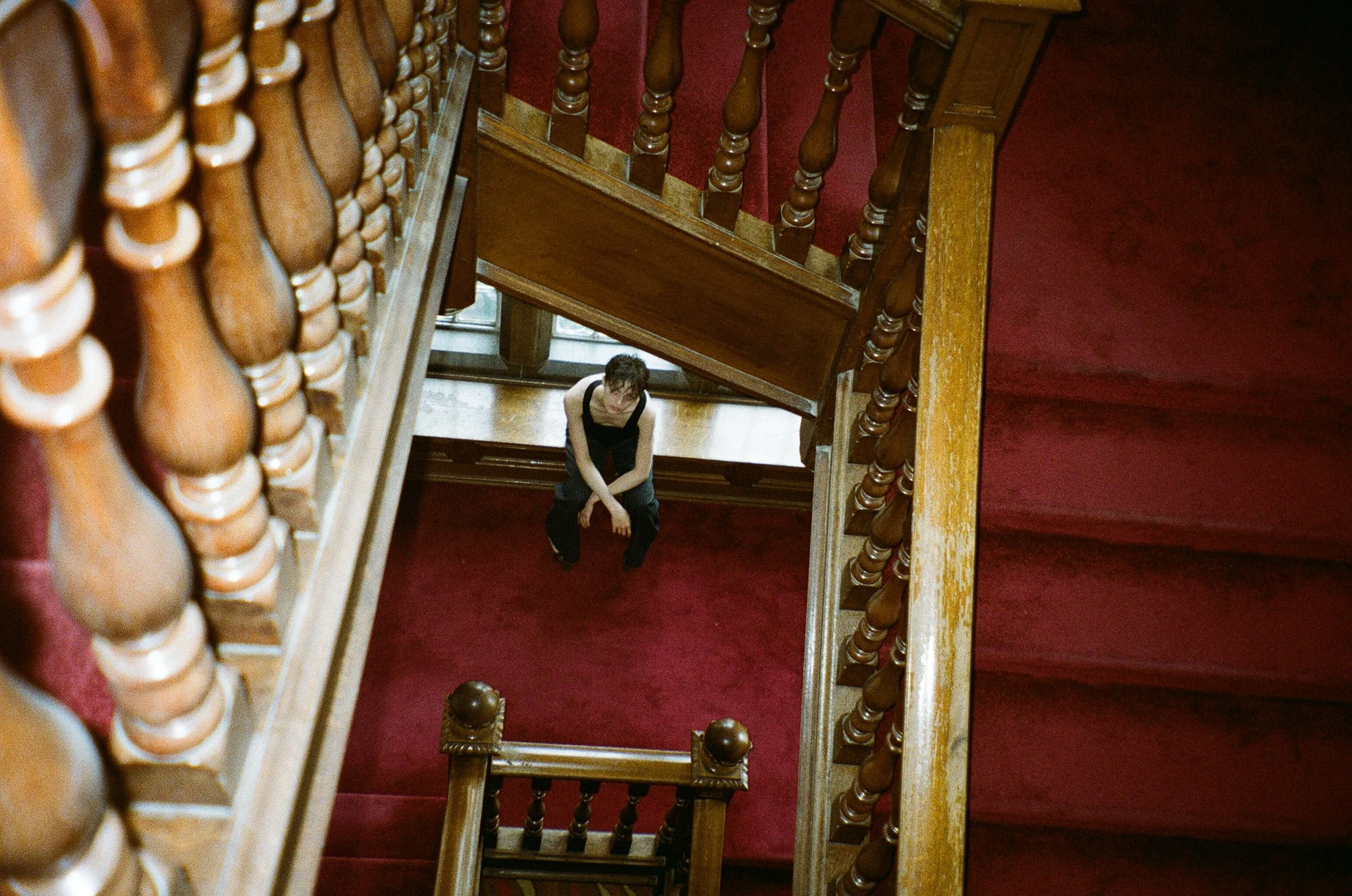 A person sitting on a wooden staircase with red carpet, viewed from above, surrounded by wooden balustrades.