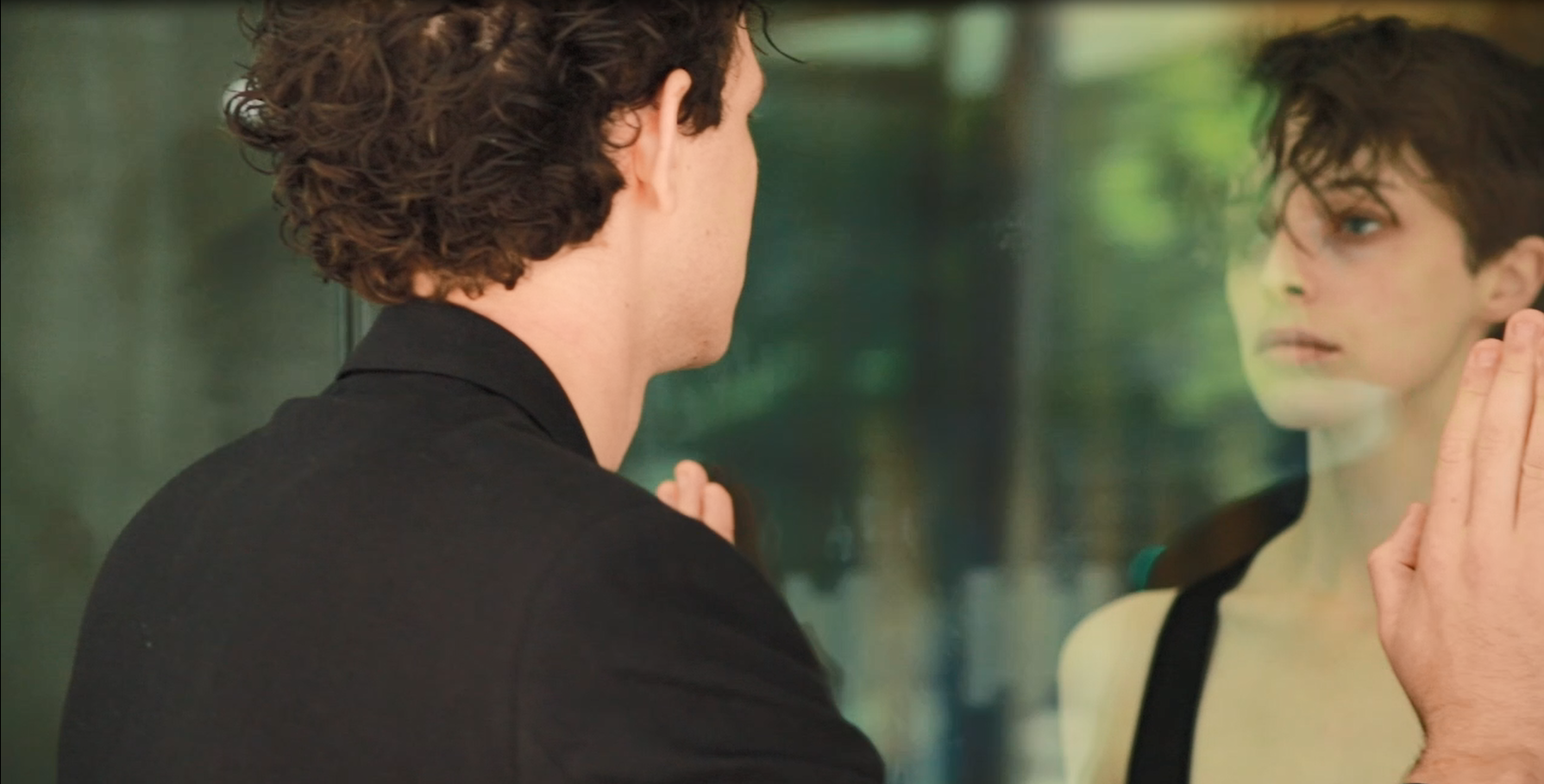 A man with curly hair is touching a glass surface while looking at a woman with short hair and blue eyes on the other side of the glass, with greenery in the background.