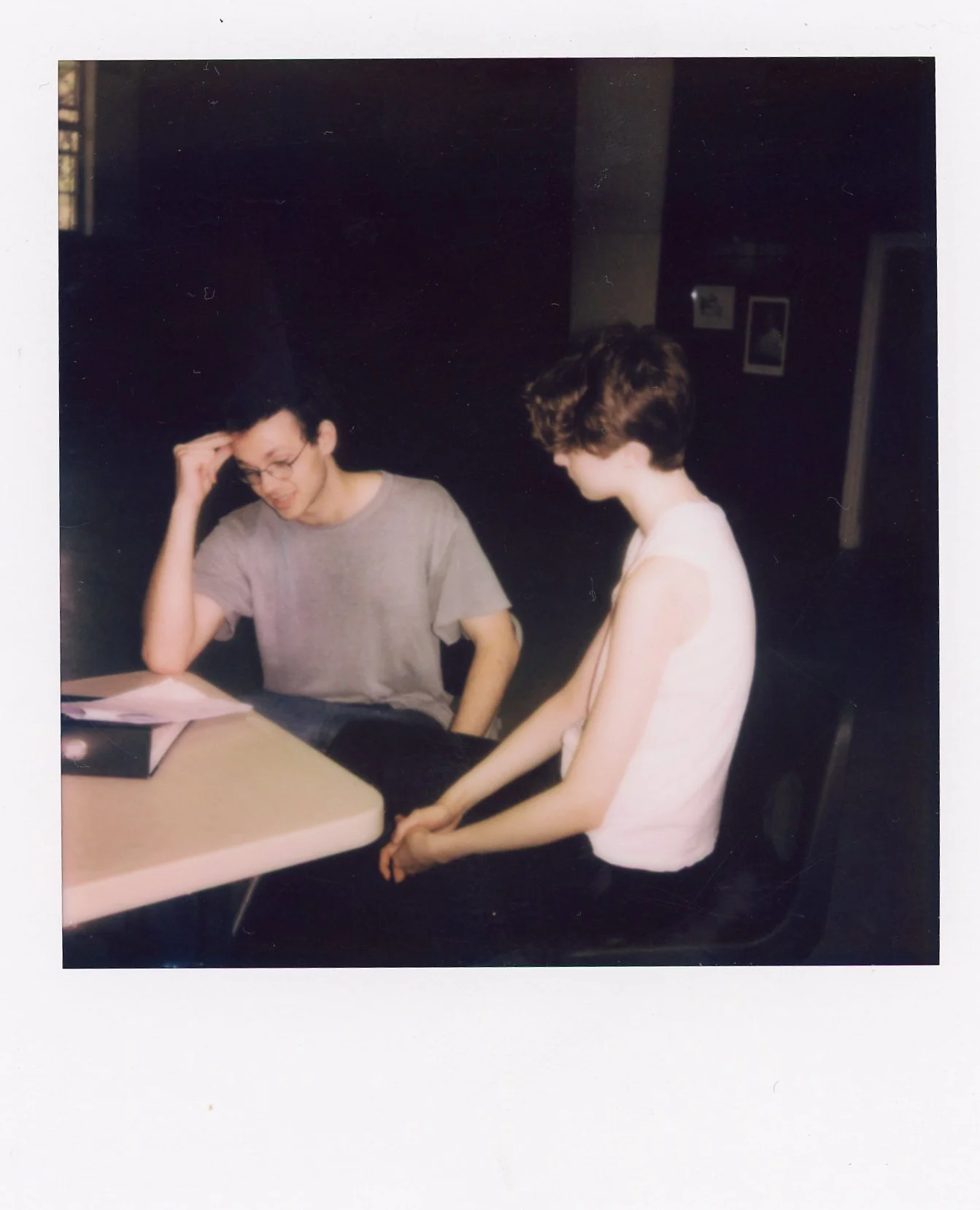 Two young people sitting at a table indoors, one leaning on their hand and the other with their hands clasped, appearing to be engaged in a conversation.