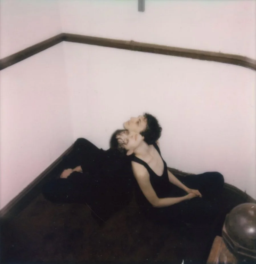 Two women with black hair and black dresses sitting on the floor in a corner with white walls and a wooden railing.