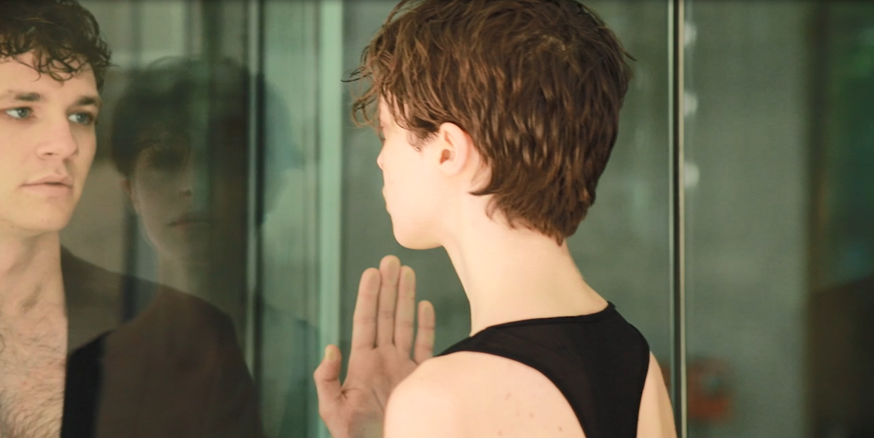 A person with short brown hair and a sleeveless black top touching a glass window, looking at their reflection.