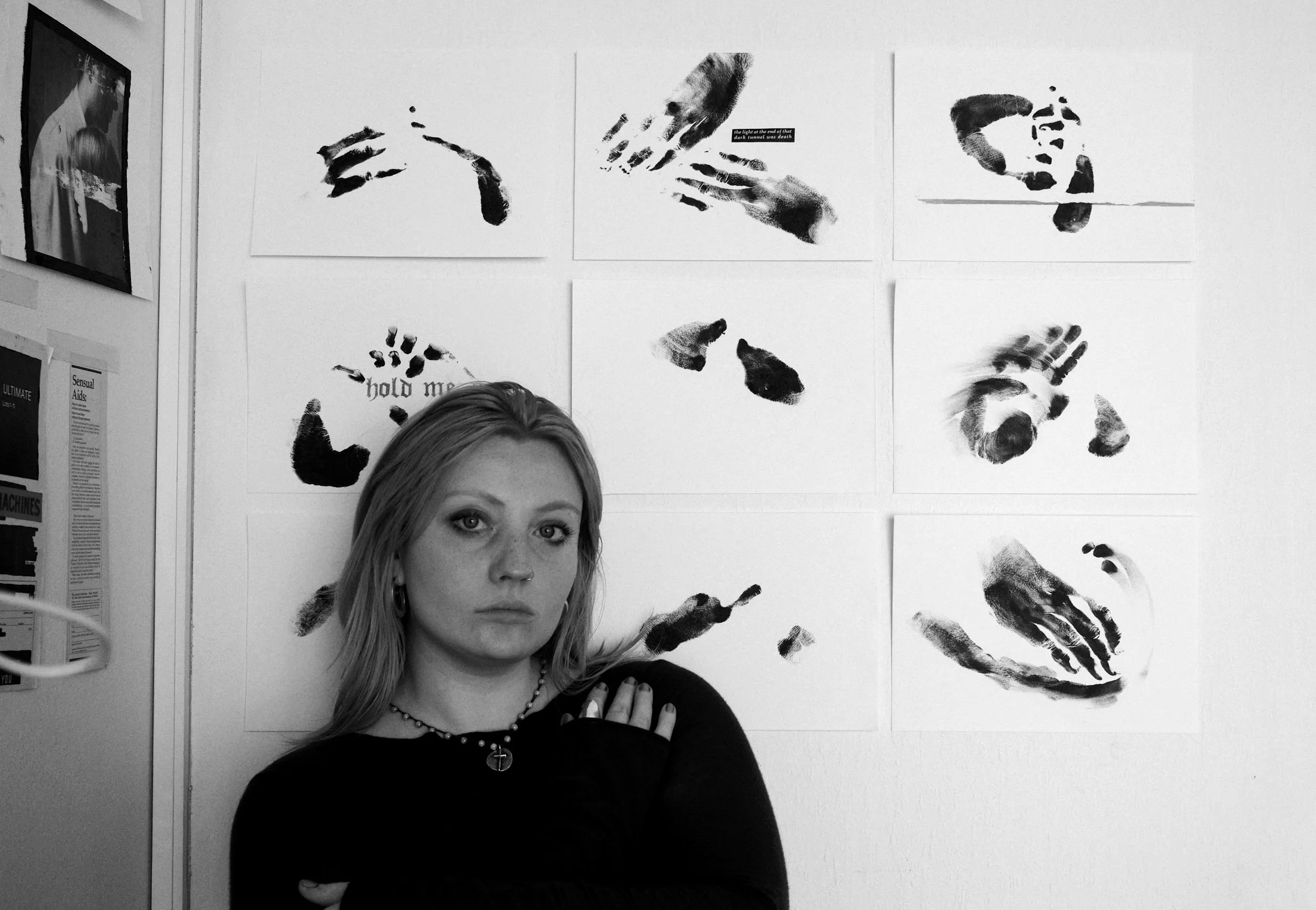A woman with long hair and a serious expression, wearing a dark shirt, is seated in front of eight portraits of handprints, some with text, displayed on a white wall.