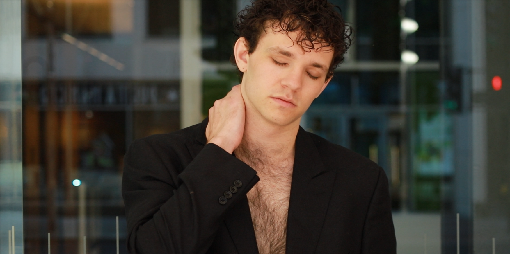 Man with curly hair and light skin, wearing a black blazer with an open shirt, touching his neck with eyes closed inside a modern building with glass doors and windows.