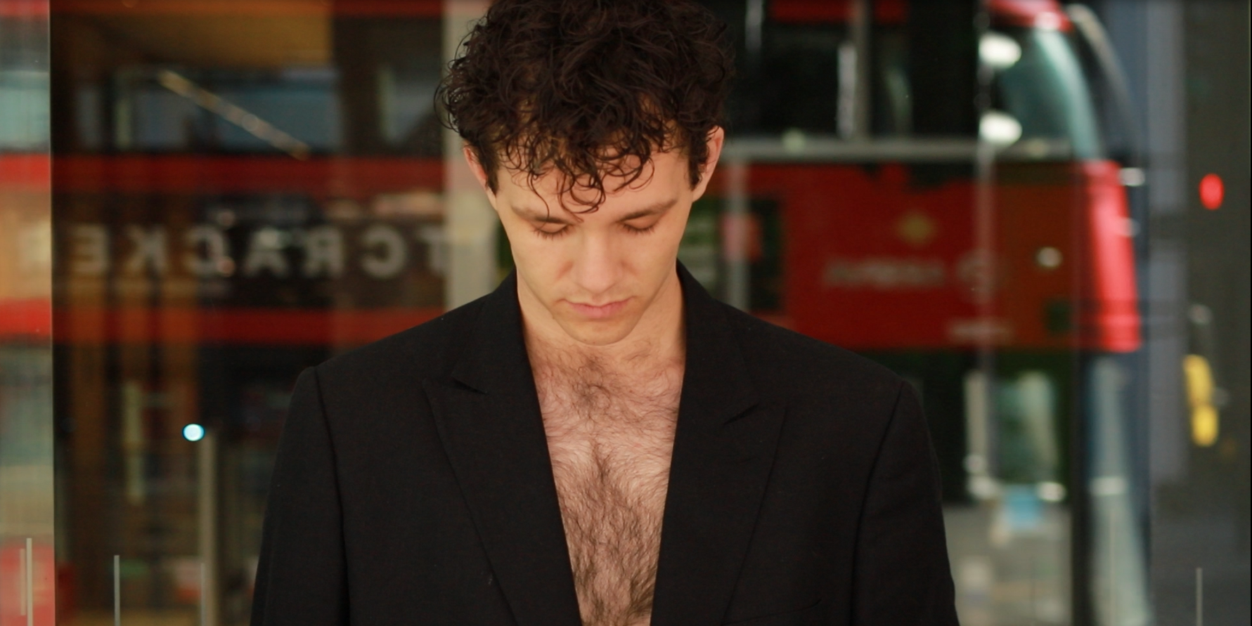A young man with curly brown hair looking down, wearing a black jacket with an open chest, standing indoors with a blurred red and green background.