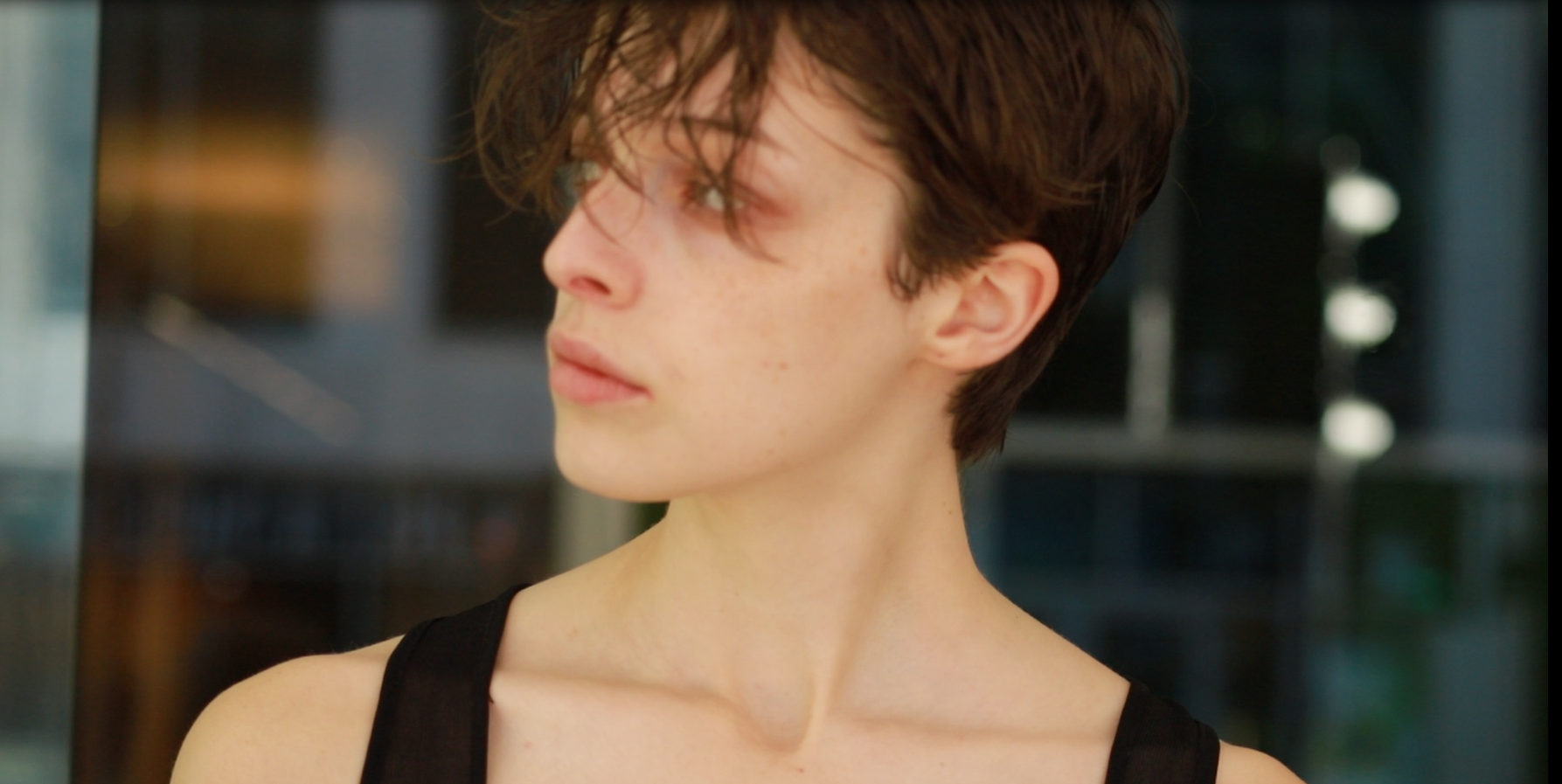 A woman with short messy brown hair and fair skin looking to the left, wearing a black sleeveless top, with a blurred modern building background.