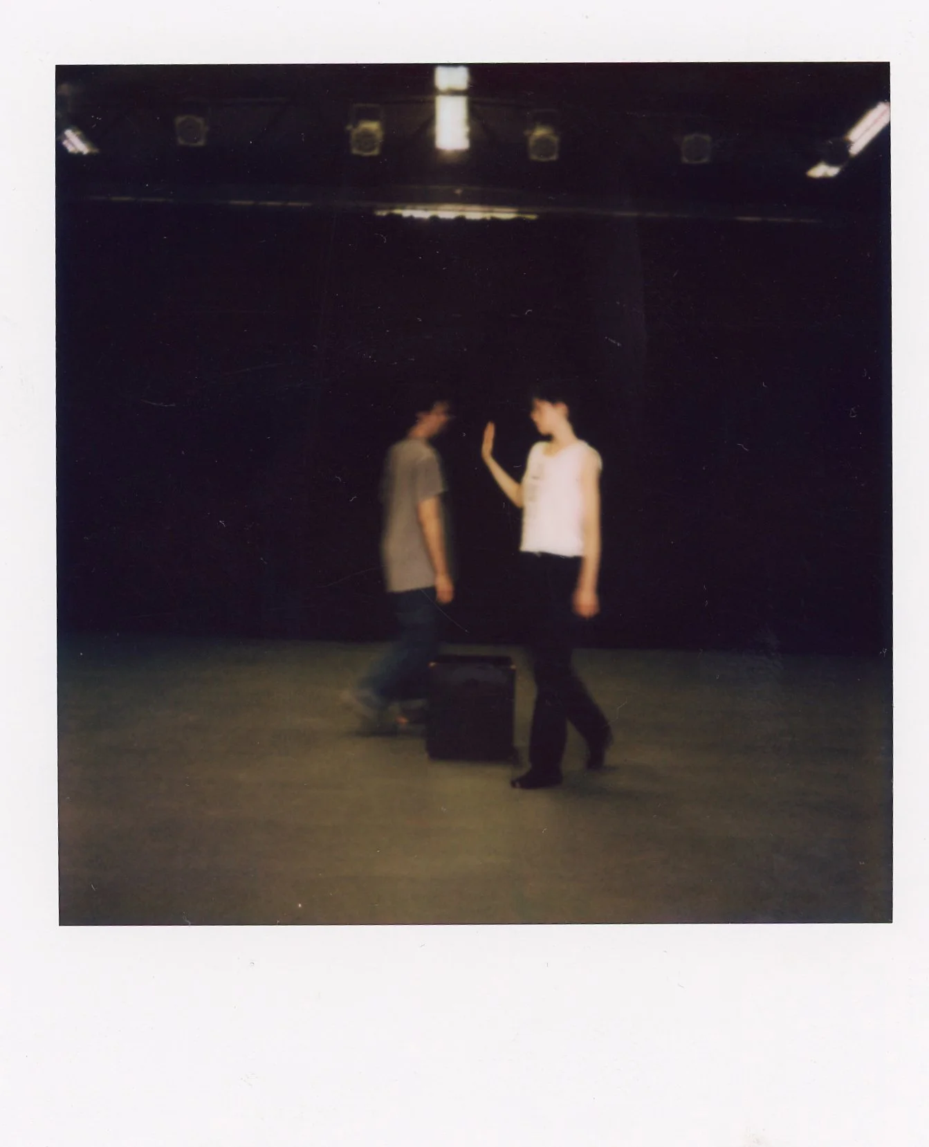 Two people standing on stage, one with a hand raised, with a speaker box between them, in a dark setting with stage lights overhead.