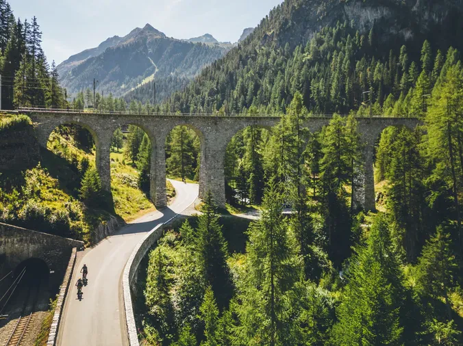 A scenic mountain landscape featuring a stone arch bridge over a winding road amidst dense green pine forests, with mountain peaks in the background under a clear blue sky.