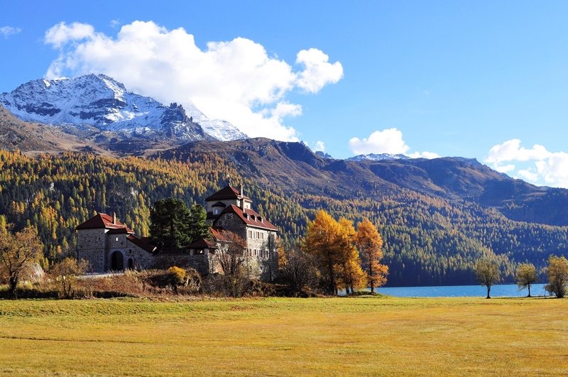 A scenic landscape featuring an old stone castle with red roofs in a grassy field, surrounded by autumn-colored trees, with a mountain range topped with snow in the background and a blue sky with white clouds.