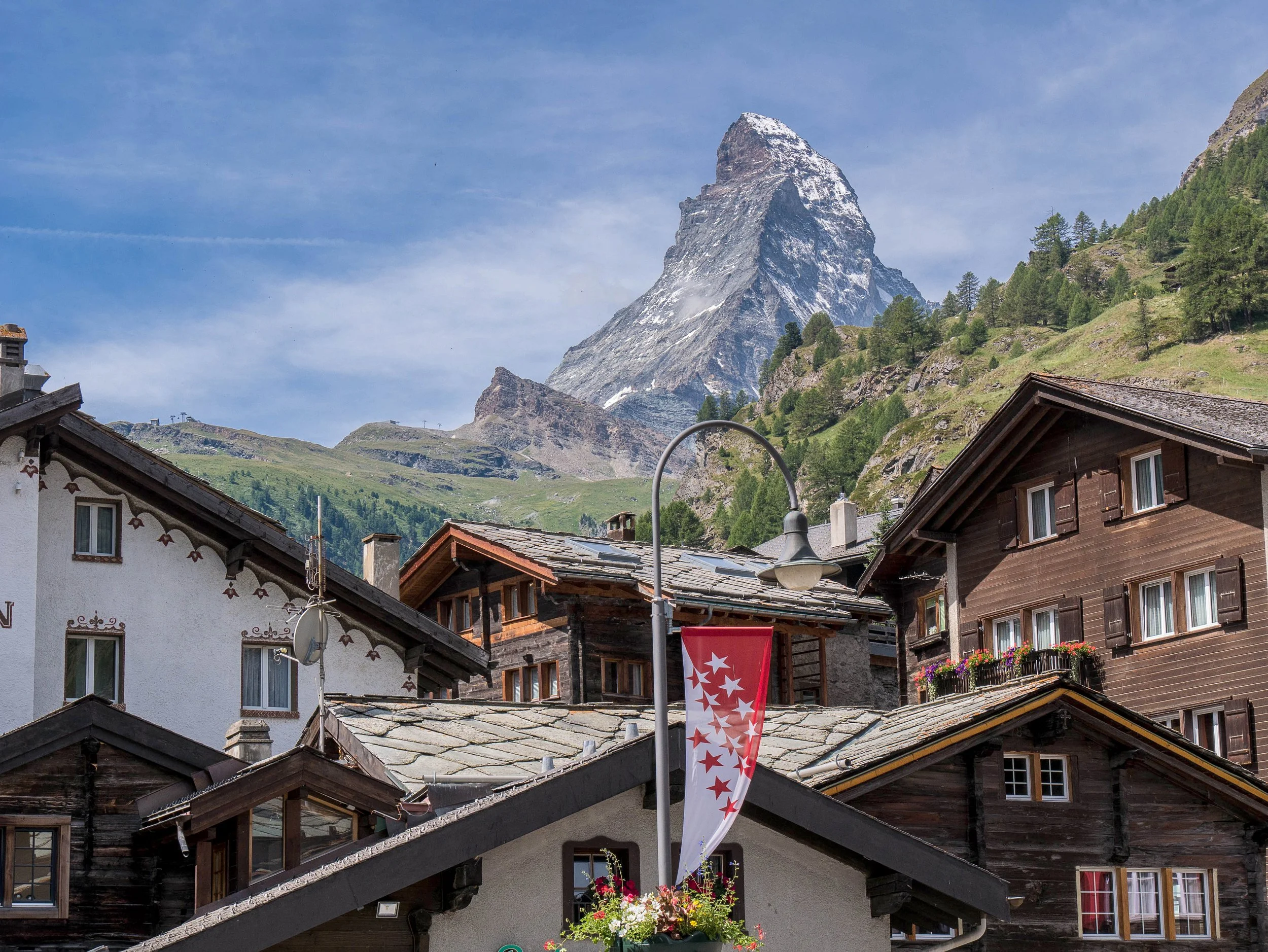 Mountain landscape with rocky peak and green hillside, Swiss village with traditional wooden and stone houses, and a Swiss flag hanging among the rooftops.