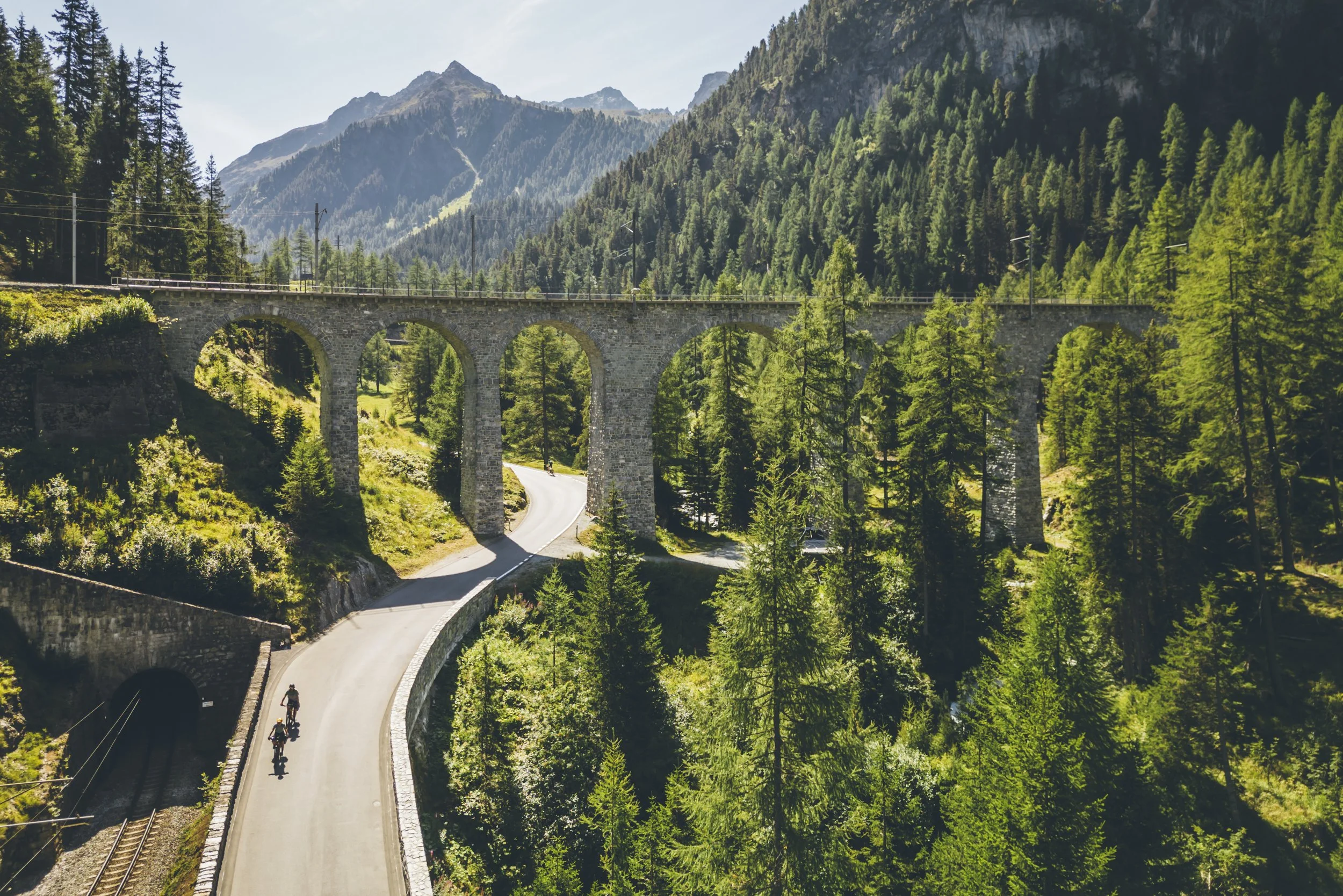 A scenic mountain landscape with a stone railway viaduct crossing a lush green forest, with a winding road passing underneath. Two cyclists are riding on the road.