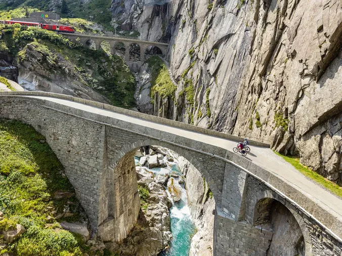 A cyclist riding a bike on a narrow mountain road with a stone bridge over a rocky river below, surrounded by steep cliffs.