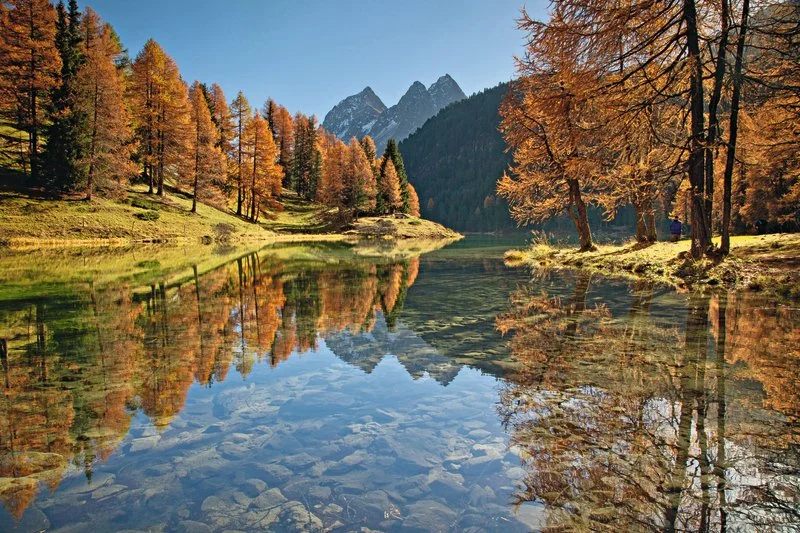 A serene mountain landscape featuring a calm lake reflecting colorful fall trees and towering mountain peaks in the background.