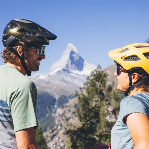 Two people wearing helmets and sunglasses outdoors with mountains and a snowy peak in the background.