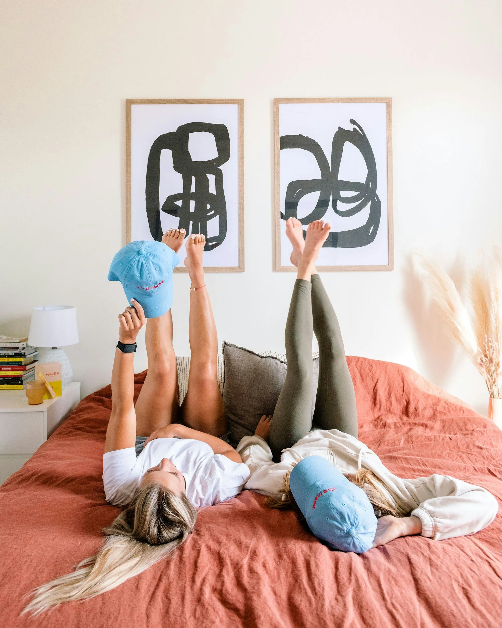 Two women lying on a bed with their legs in the air, one holding a blue cap, in front of two abstract black and white artworks on the wall.