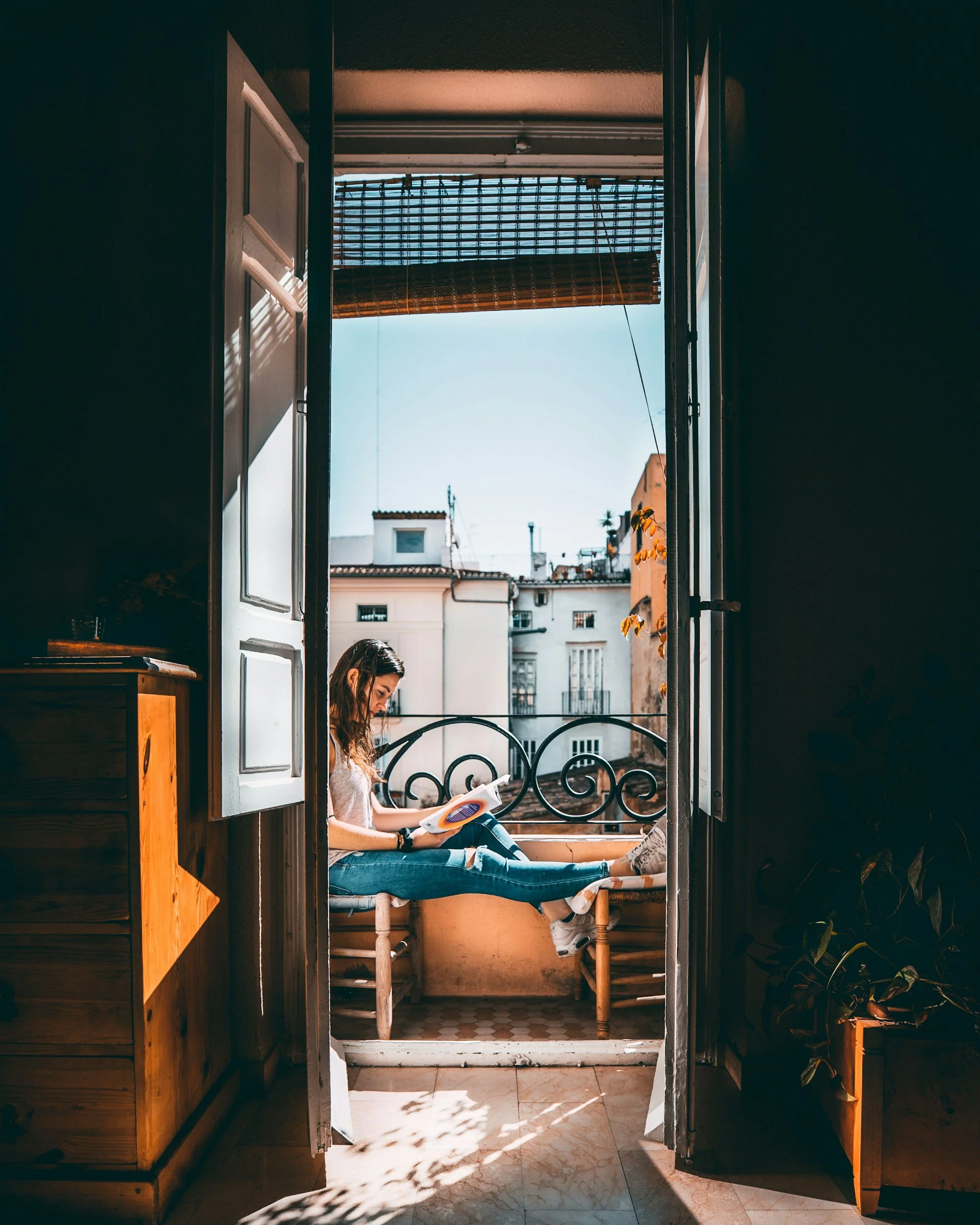 A young woman sitting on a balcony, reading a book during daytime, with buildings and a clear sky in the background.