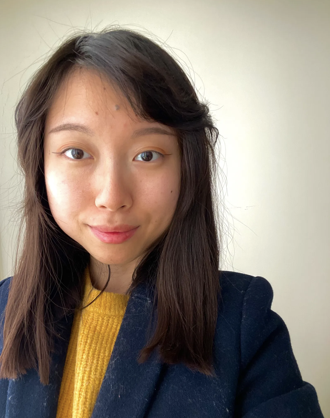 A young woman with shoulder-length dark hair, wearing a yellow sweater and a dark blazer, smiling at the camera indoors.