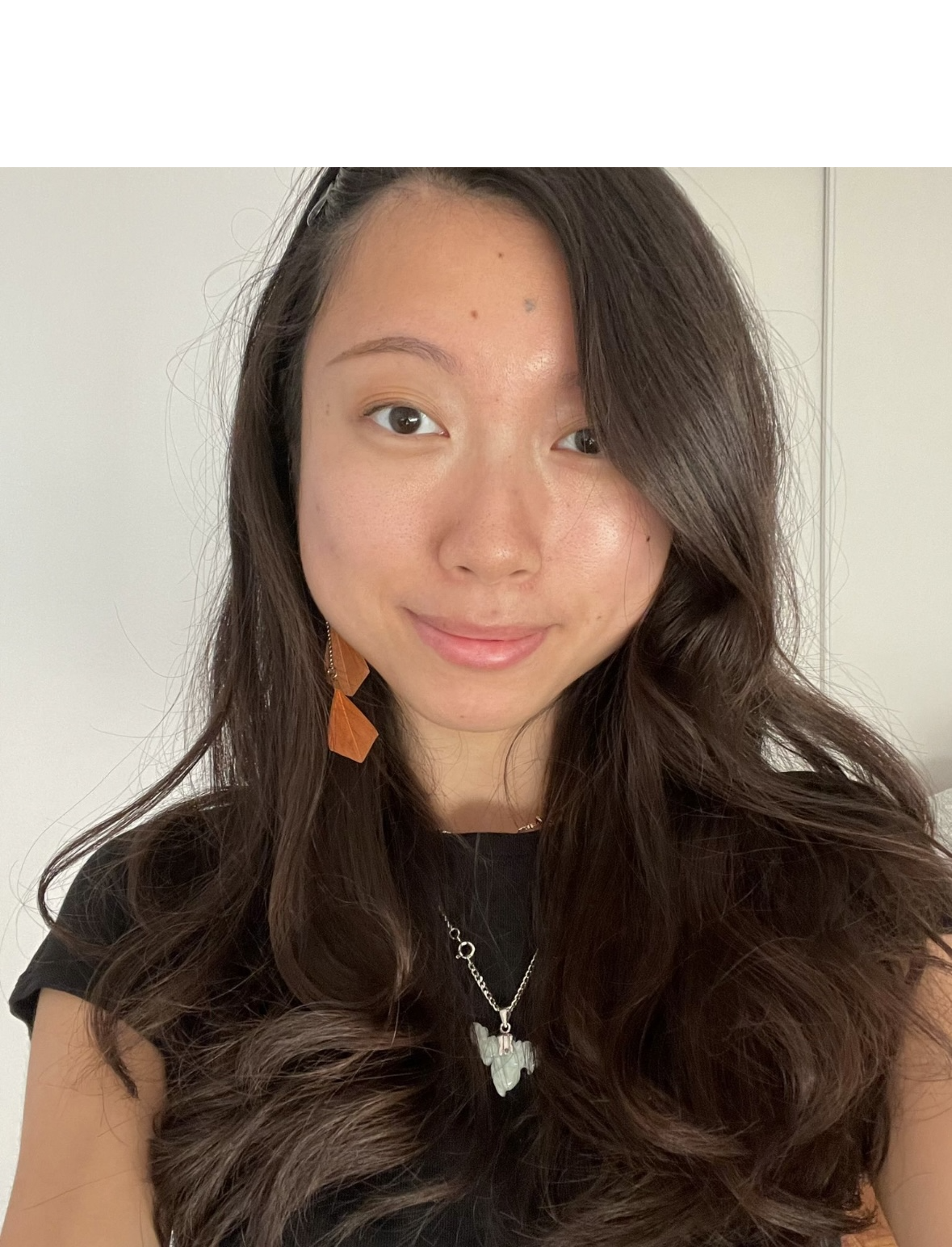 Close-up of a young woman with long wavy dark hair, wearing orange earrings, a butterfly necklace, and a black top, smiling softly.
