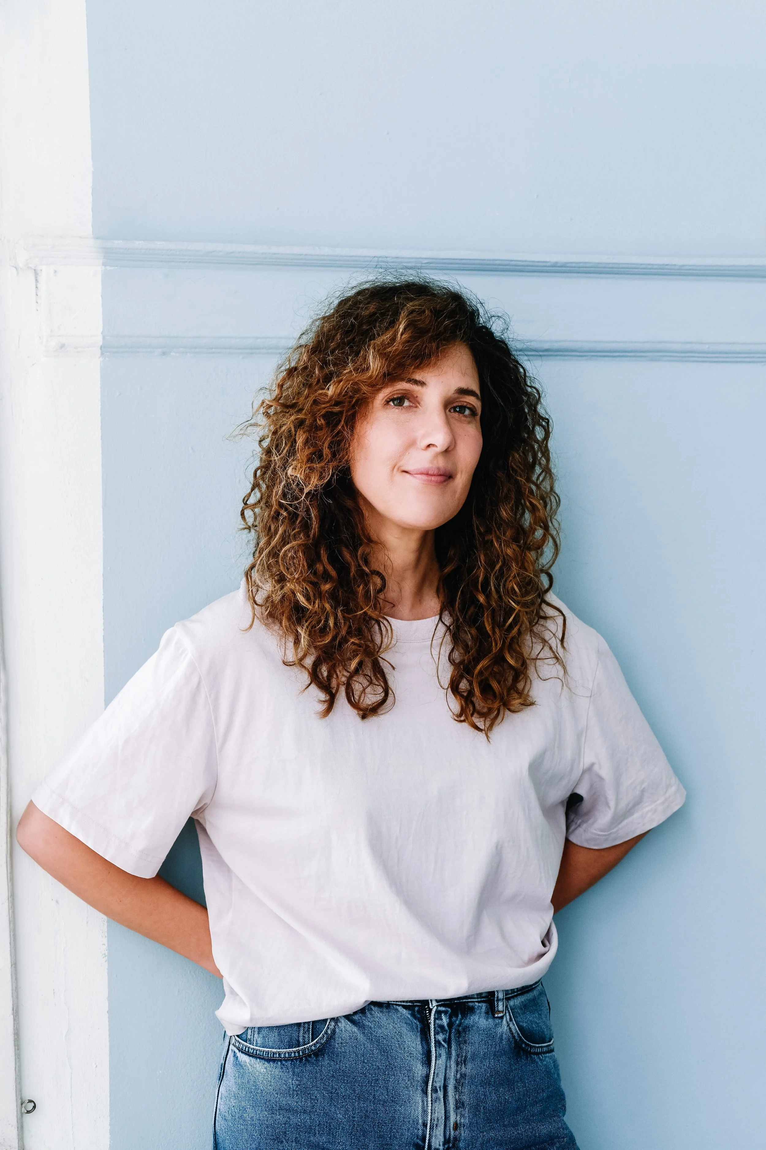 A woman with curly brown hair wearing a white t-shirt and blue jeans, standing with her hands behind her back against a light blue wall.