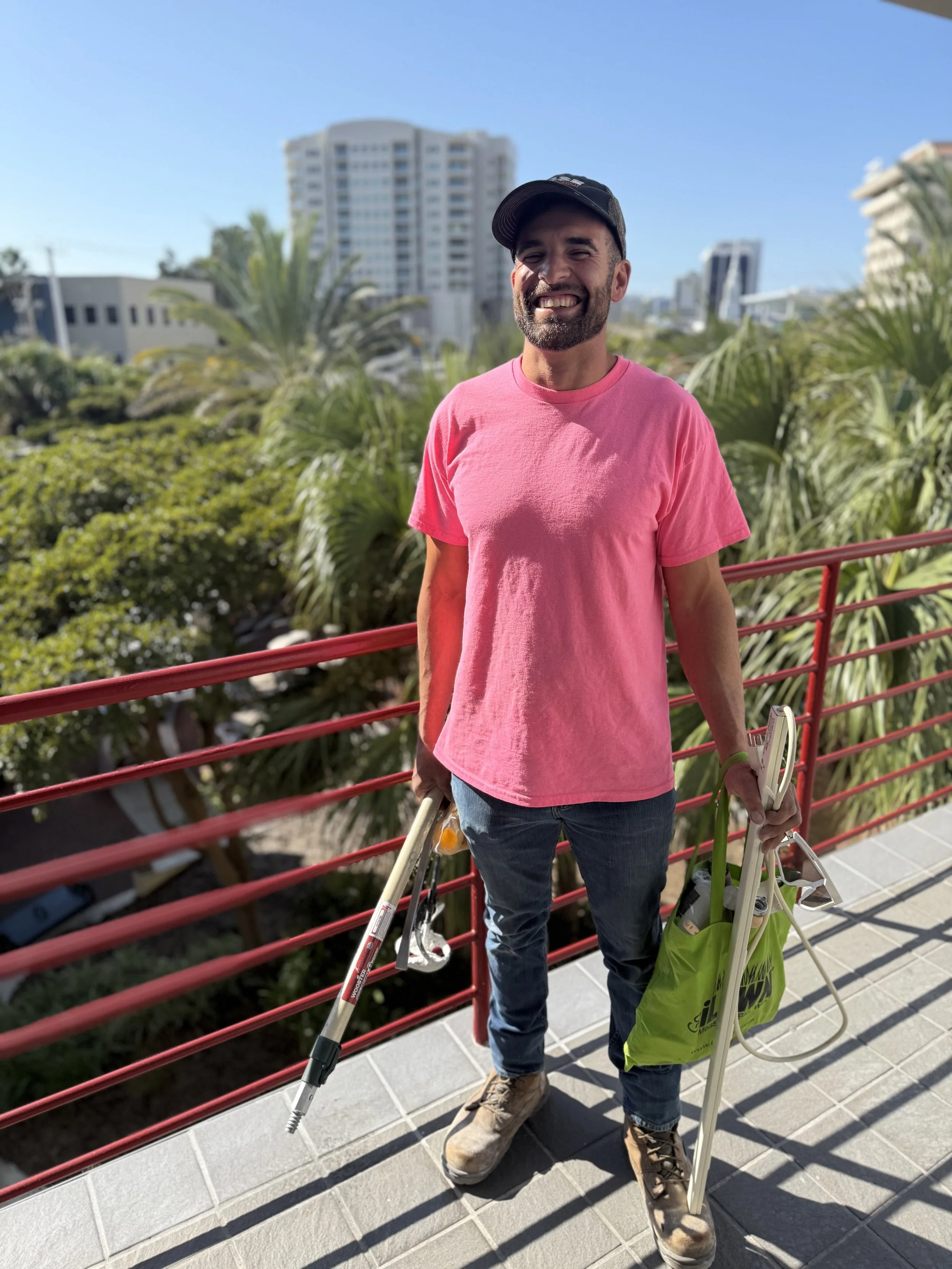 Owner of Pinky's with a beard smiling, wearing a pink t-shirt, jeans, and a black cap, stands outdoors on a balcony with a red railing, holding landscaping equipment in Sarasota.