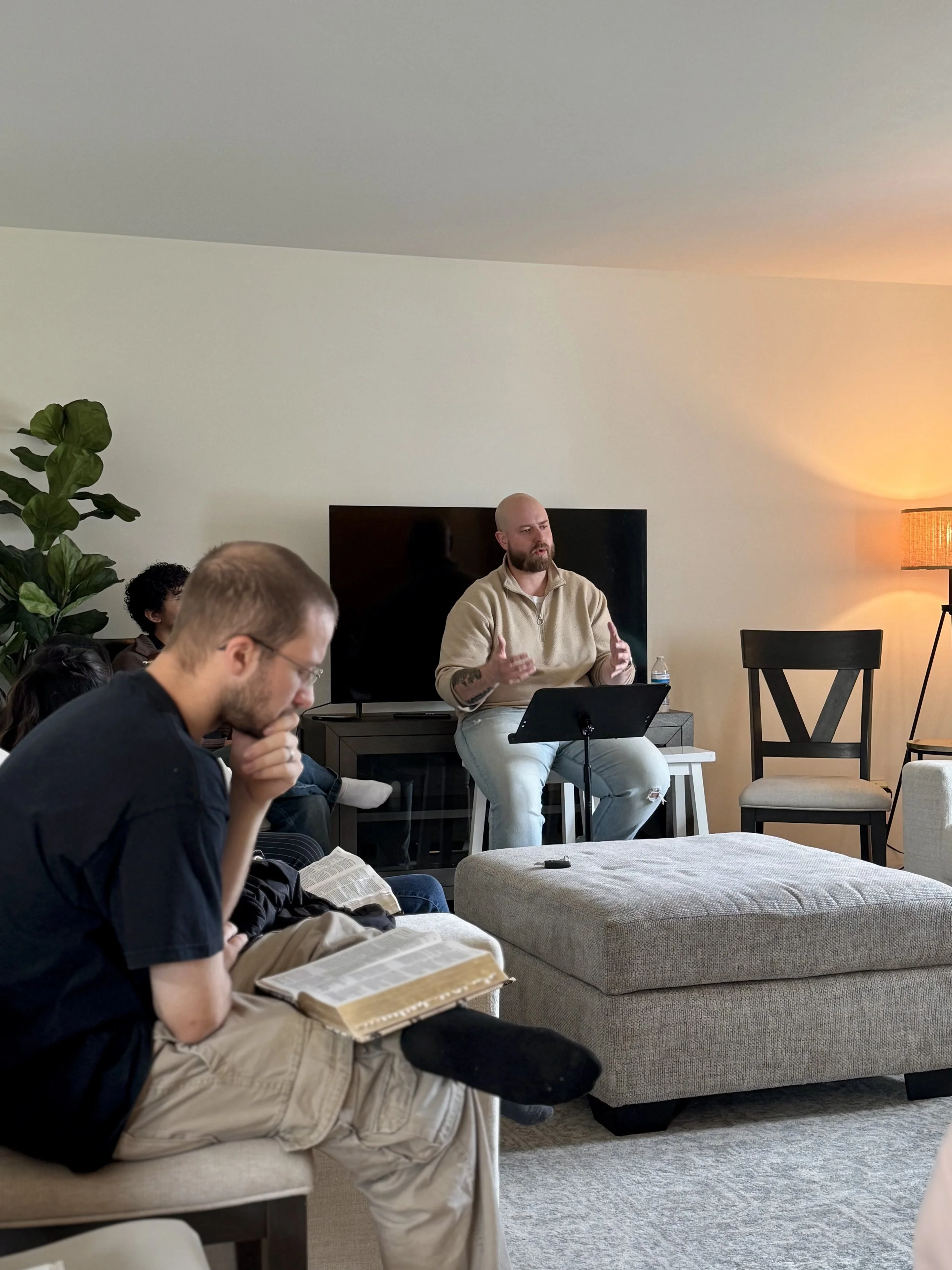 A group of people attending a meeting or discussion in a living room. One person is standing and speaking, while others are seated and listening, with some reading a book.