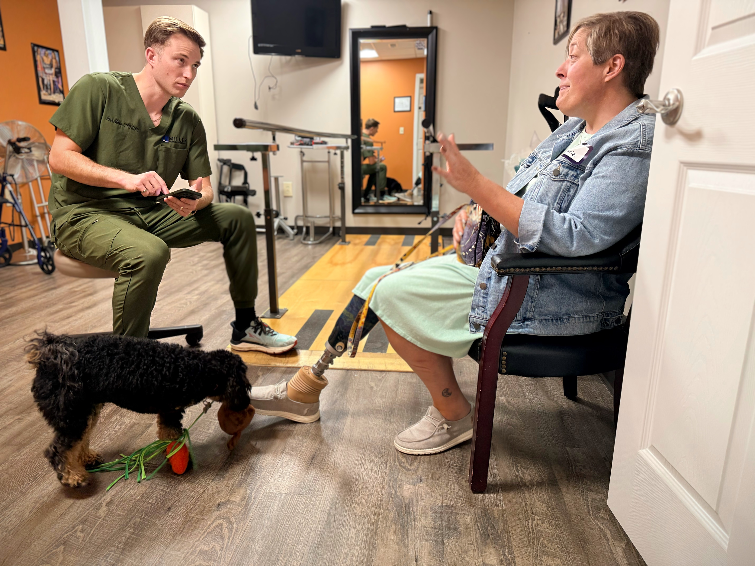 A healthcare professional in a green uniform is sitting and talking to a woman with a prosthetic leg, sitting in a chair, wearing a denim jacket and a name tag. The woman's prosthetic leg is a new custom-designed limb. A therapy dog with a toy is wal