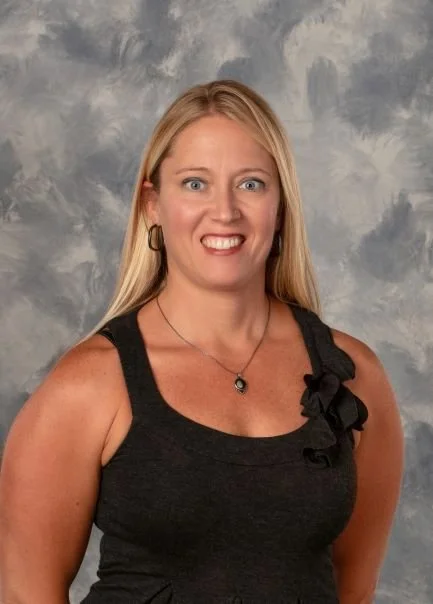 A woman with long blonde hair wearing a black dress with a ruffle detail on the shoulder, gold hoop earrings, and a necklace with a black pendant, smiling against a cloudy gray background.