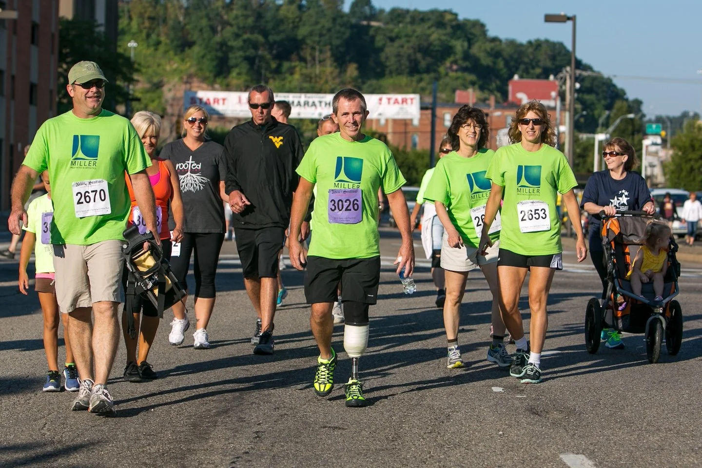 Group of people participating in a marathon or race, wearing colorful athletic clothing and race bibs, with some pushing strollers, walking on a city street.