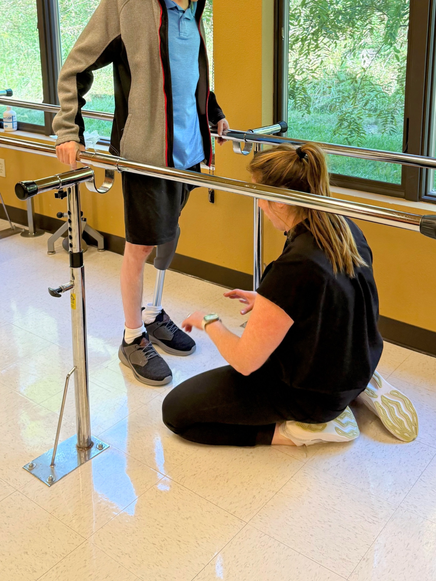 Young man with a prosthetic leg, standing with the support of parallel bars, being assisted by a woman kneeling on the floor, in a physical therapy or rehab setting.
