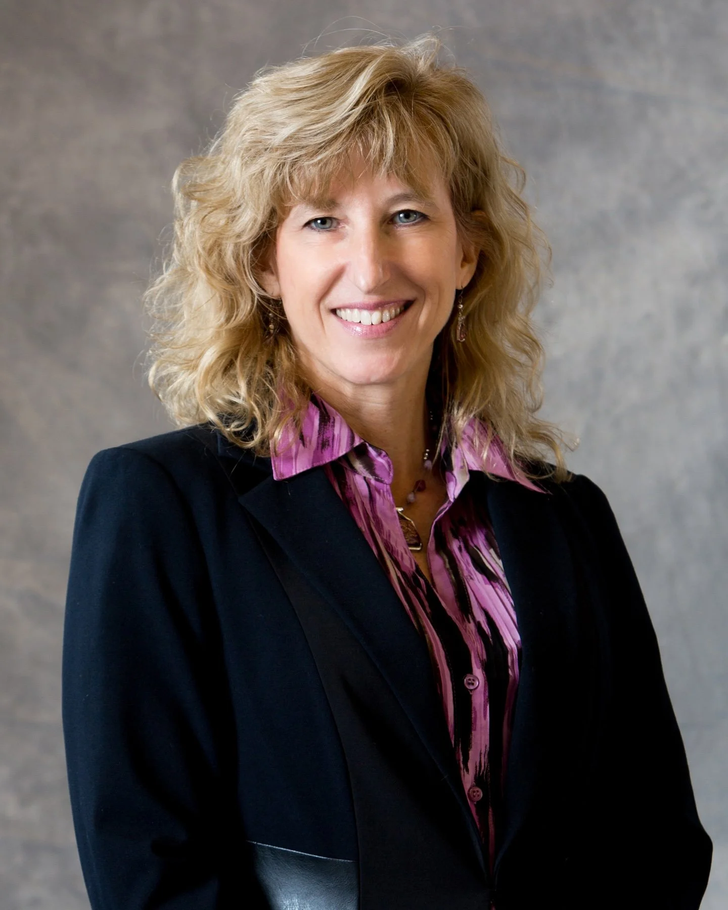 A middle-aged woman with blonde, curly hair, smiling, dressed in a black blazer over a purple and pink striped shirt, standing against a neutral background.