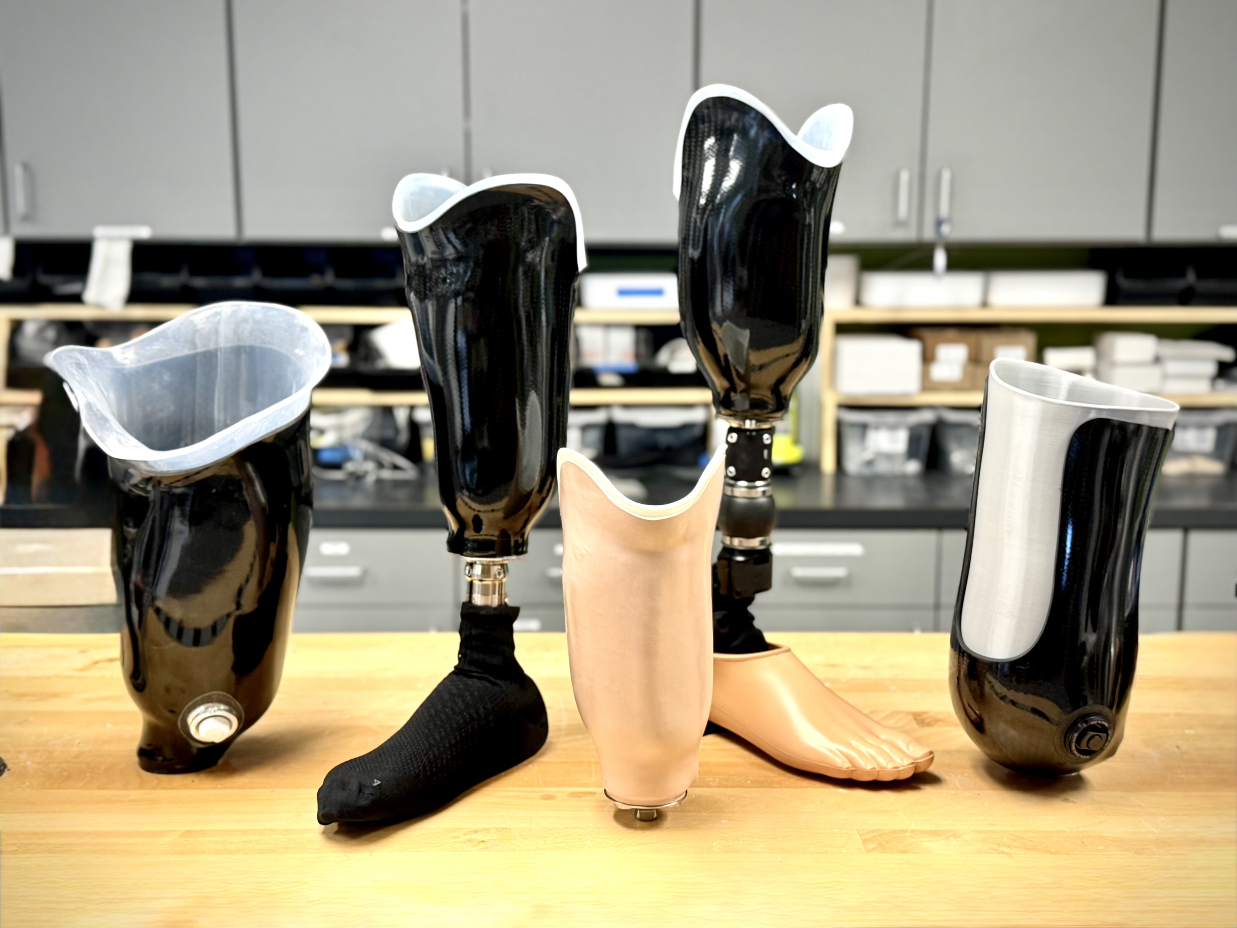 Five prosthetic limbs arranged on a wooden table in a workshop.
