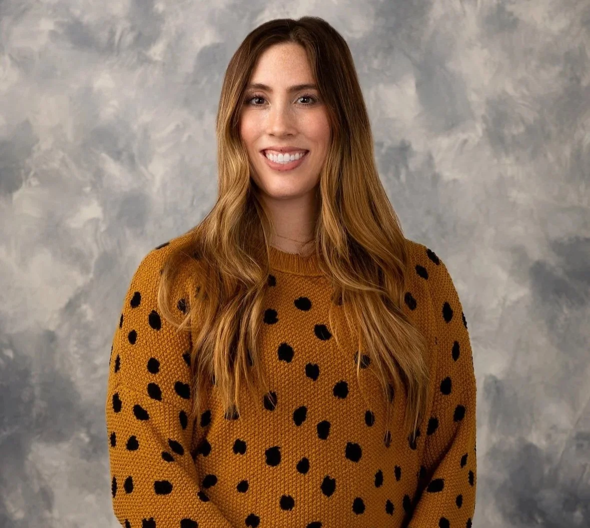 A woman with long, wavy red hair smiling at the camera, wearing a mustard yellow sweater with black polka dots, against a gray cloud-like background.