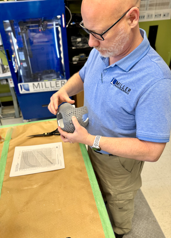 A man holding a gray 3D-printed prosthetic hand in a workshop or laboratory setting with equipment and a blue machine in the background.