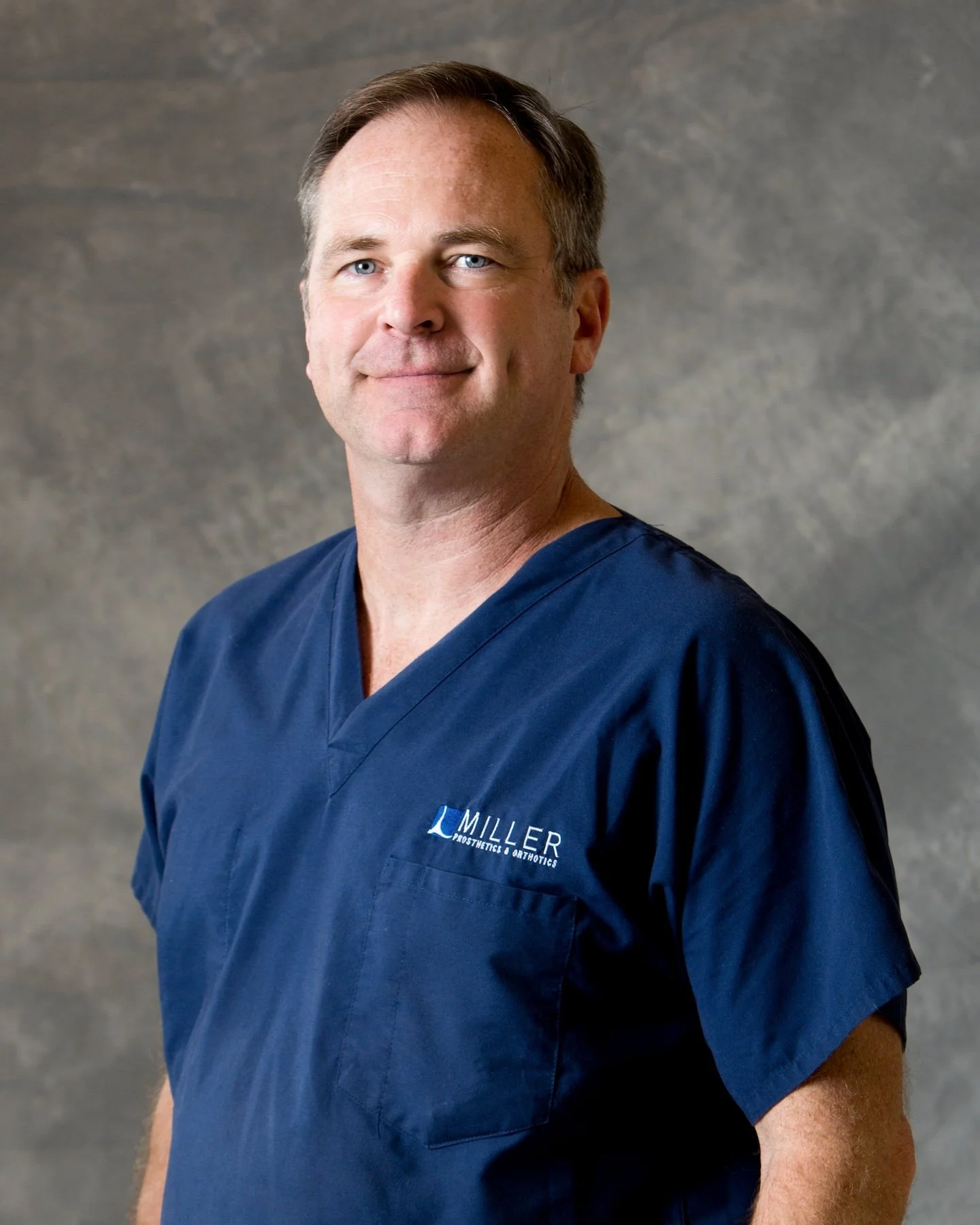 Portrait of a male healthcare professional wearing blue scrubs with a logo that reads 'Miller' and 'Plastic & Orthopedic Surgery' in front of a textured grey background.