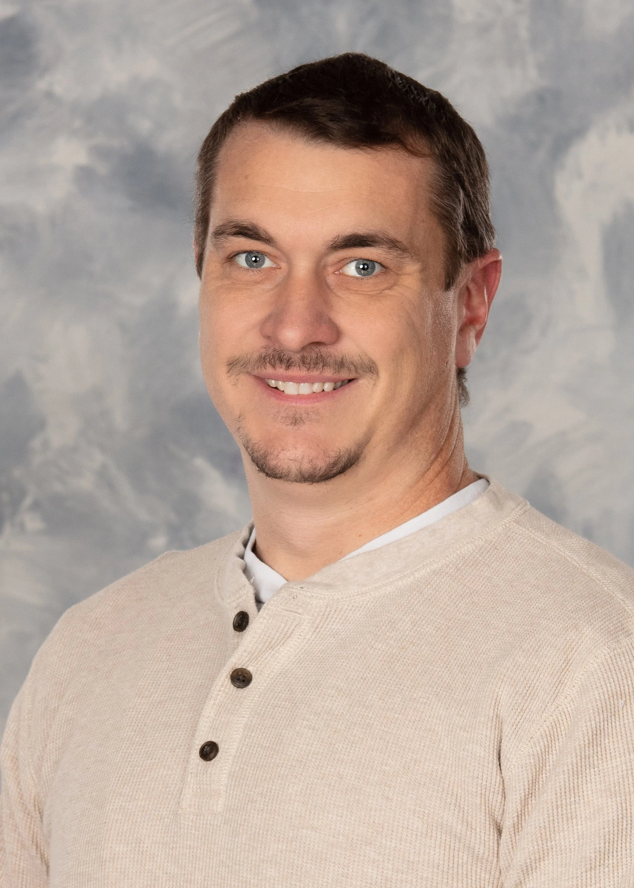 A man with short brown hair, blue eyes, and a goatee, smiling, wearing a beige shirt with buttons, against a cloudy sky background.