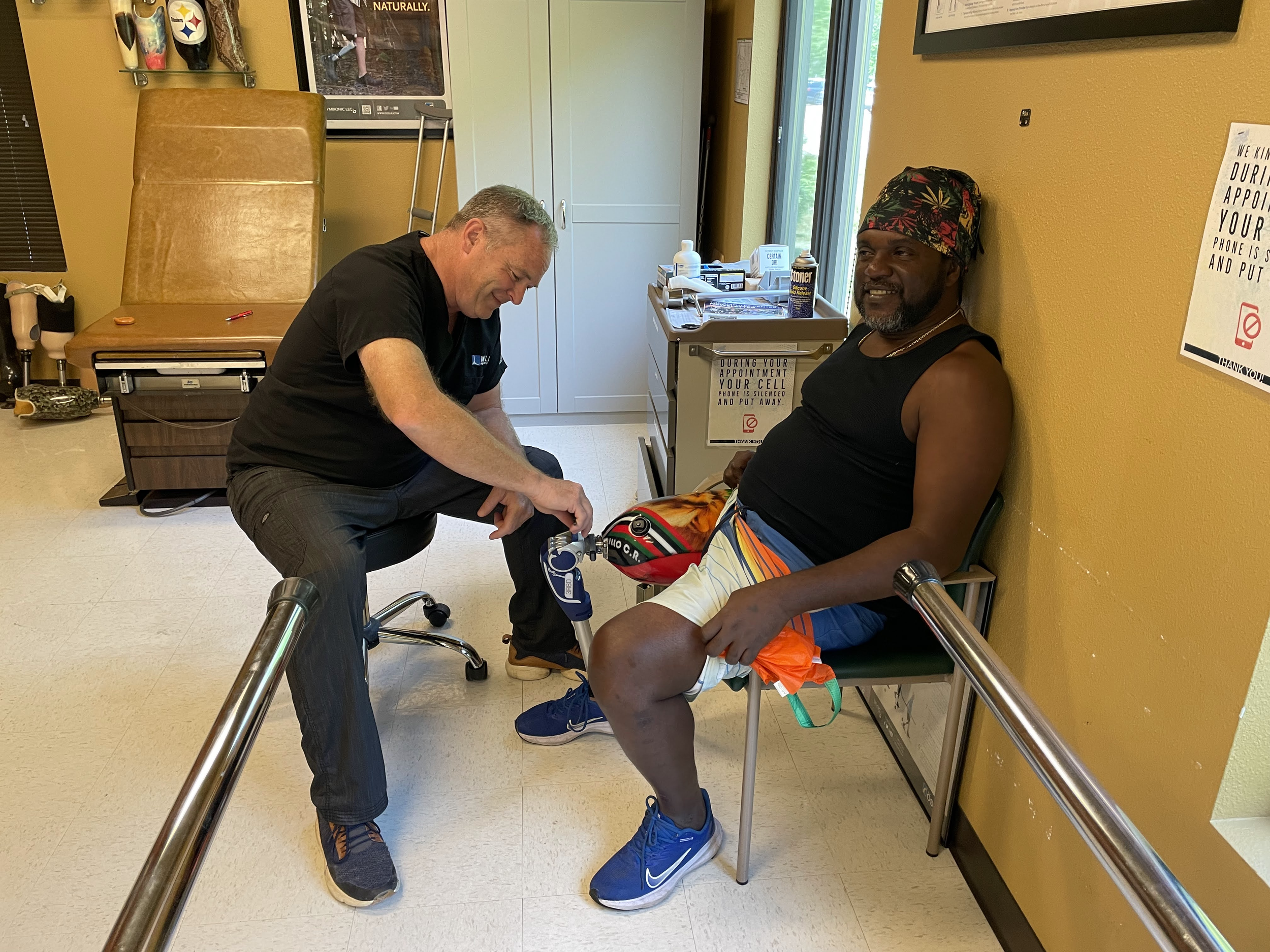 A man sitting in a medical chair at a clinic, smiling while a healthcare worker checks his leg, which is in a cast, in a room with yellow walls.