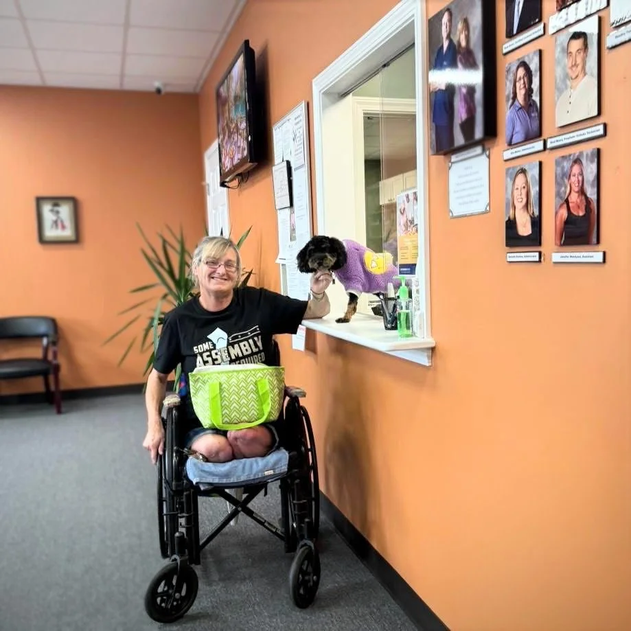 A woman in a wheelchair smiling and holding a small dog dressed in a purple sweater, with her arm extended through a window in an office or reception area. On the wall behind her are framed photographs and portraits.