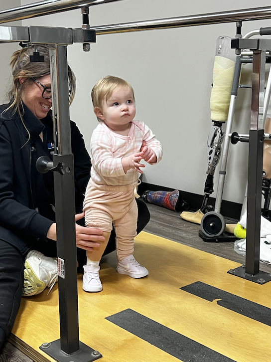 A young child standing on a yellow therapy mat, supported by an adult, with a prosthetic leg and crutches nearby in a physical therapy room.