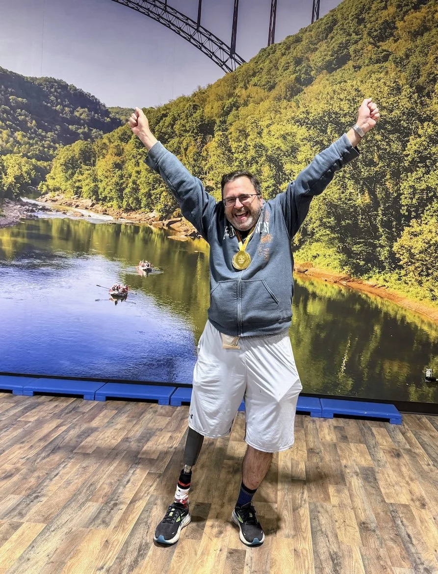 A man celebrating with arms raised, wearing a medal, standing on a wooden floor in front of a scenic outdoor backdrop of a river, trees, mountains, and a bridge, smiling broadly.