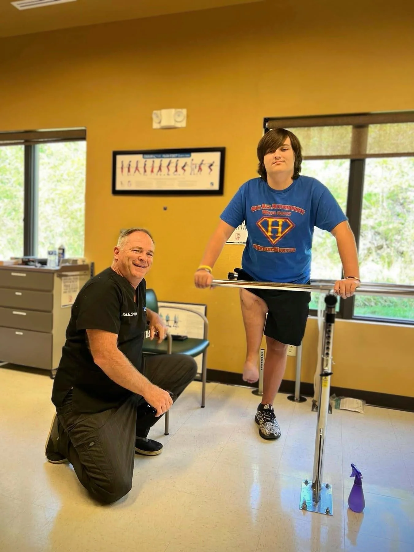 A young boy demonstrates physical therapy exercises with a bar while a therapist kneels beside him in a clinic room. The boy is balancing on one foot and holding the bar for support, wearing a blue superhero T-shirt and black shorts. The therapist is