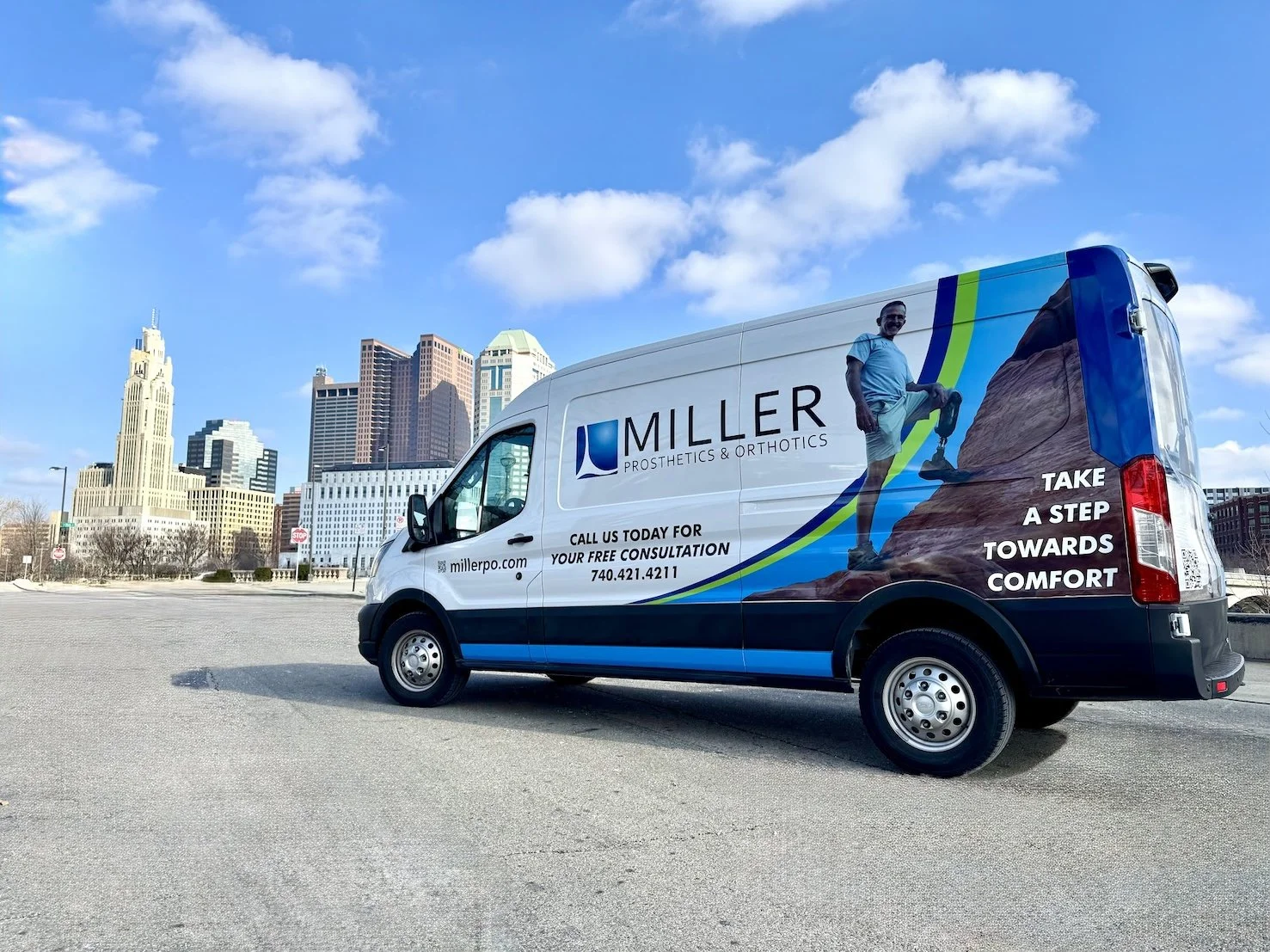 A branded van parked in an open area with a city skyline in the background. The van features a man climbing a rock wall, along with text promoting Miller Prosthetics & Orthotics, including contact information and a motivational phrase.