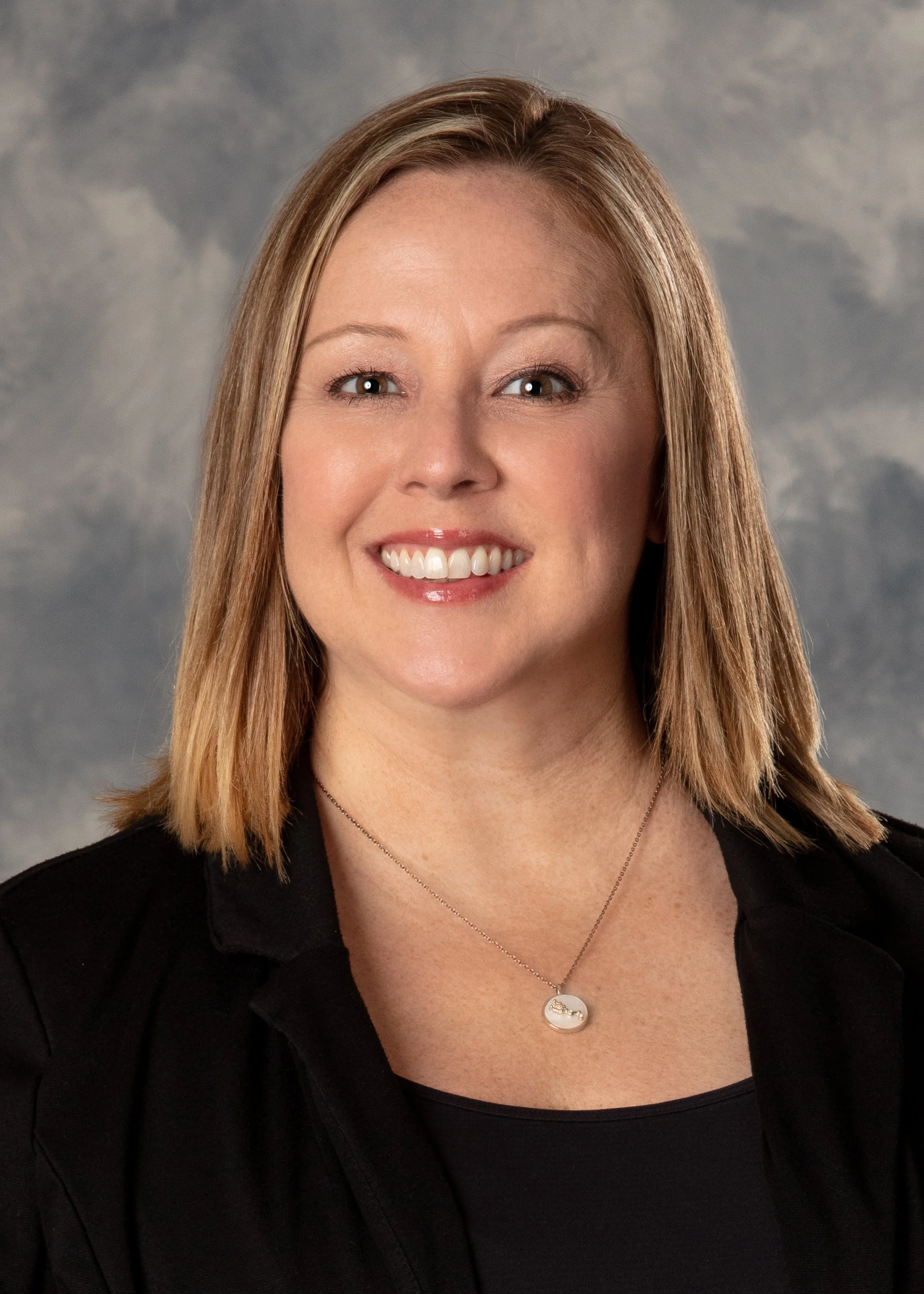 A woman with shoulder-length light brown hair smiling, wearing a black top and a silver necklace with an oval pendant, against a cloudy sky background.